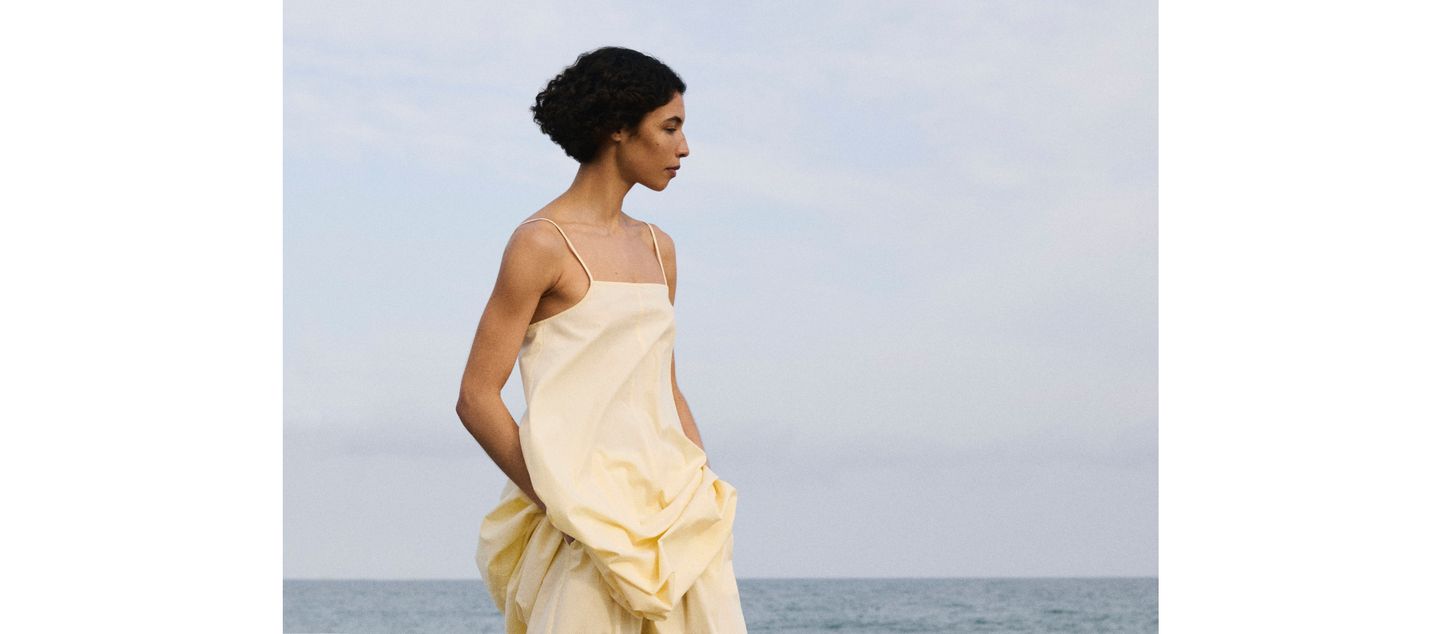 Woman in a yellow dress stands on a beach, gazing ahead with the ocean and sky in the background.