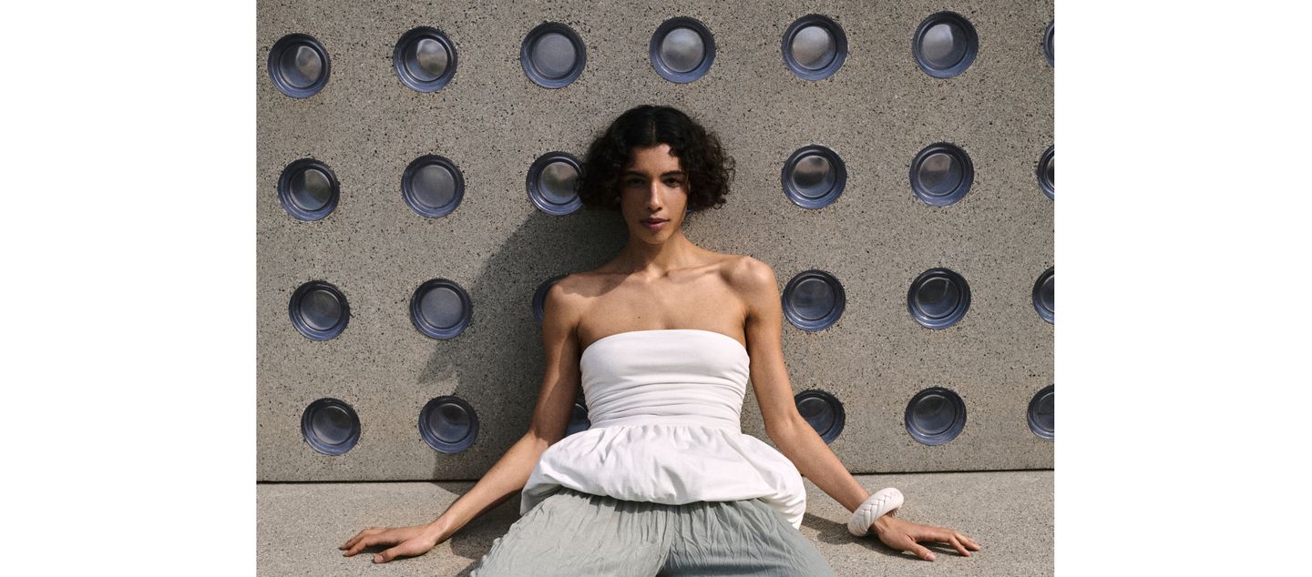 Woman in white top sits against a textured wall with circular patterns, wearing a large bracelet, in an outdoor setting.