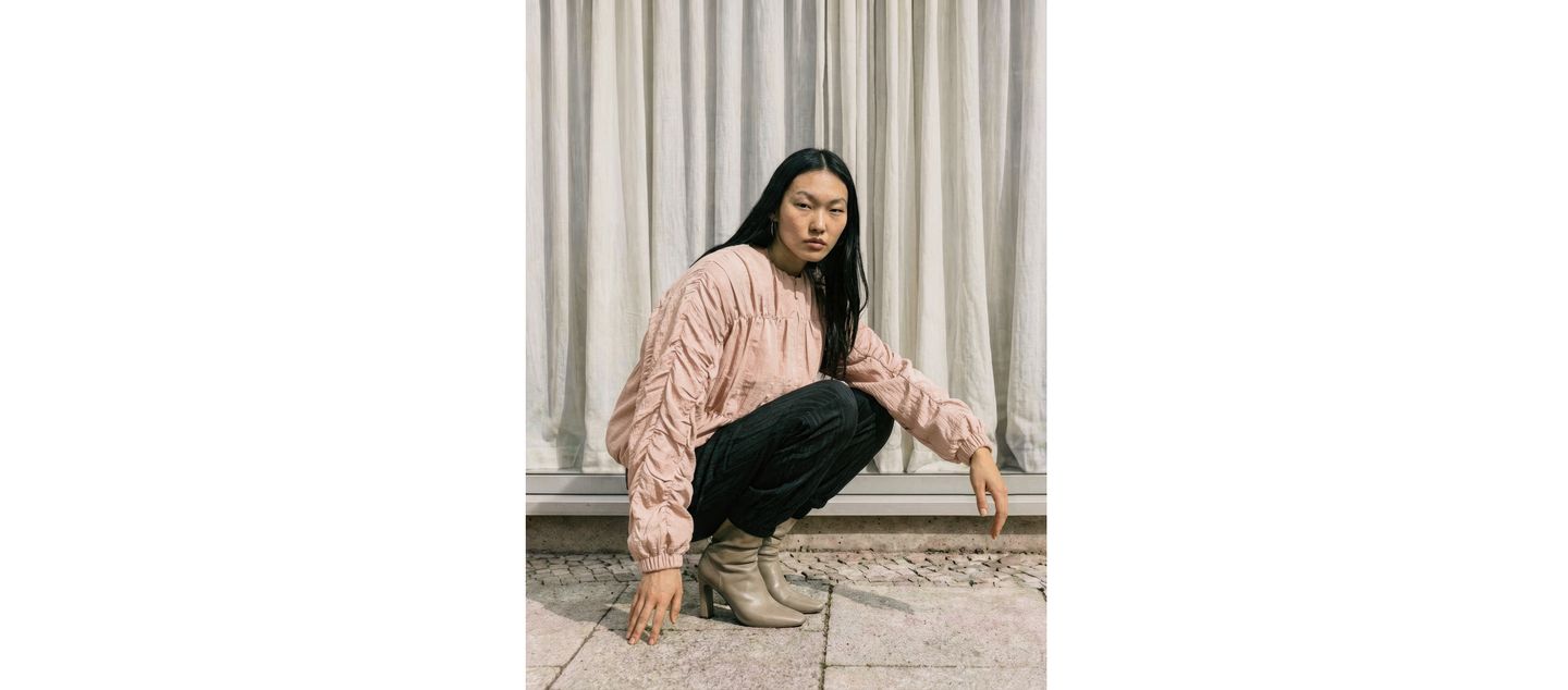 A person crouches near a window with curtains, wearing a textured pink blouse, dark pants, and light tan boots, looking at the camera.