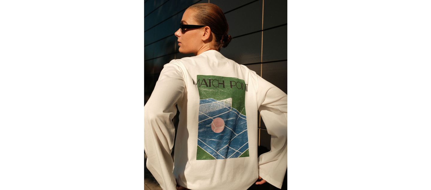 Person wearing sunglasses and a white shirt with a tennis court graphic and "MATCH POINT" text on the back, standing by a dark wall.