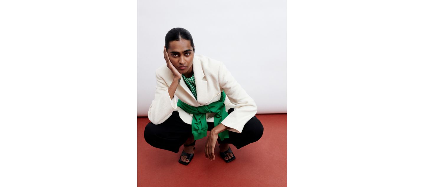 Person in a white blazer with a green belt squats on a red floor, hand on face, and wears black pants and sandals, against a plain background.