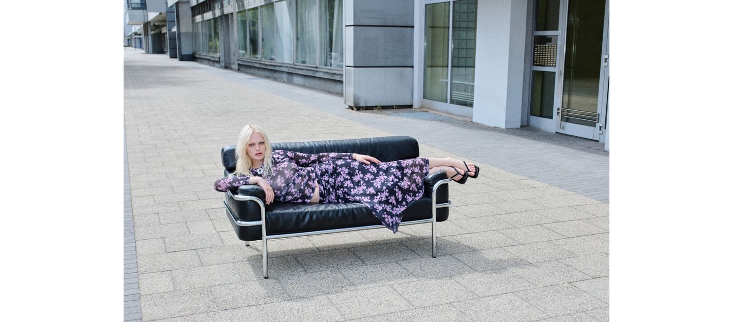 A person with long blonde hair reclines on a black leather sofa on an urban sidewalk, wearing a purple floral dress and black sandals.