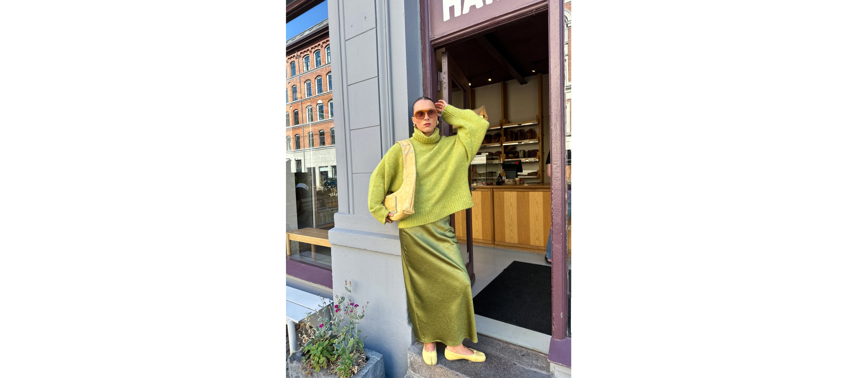 Person in a green sweater and skirt, yellow shoes, and sunglasses standing in a doorway; urban street and building in the background.