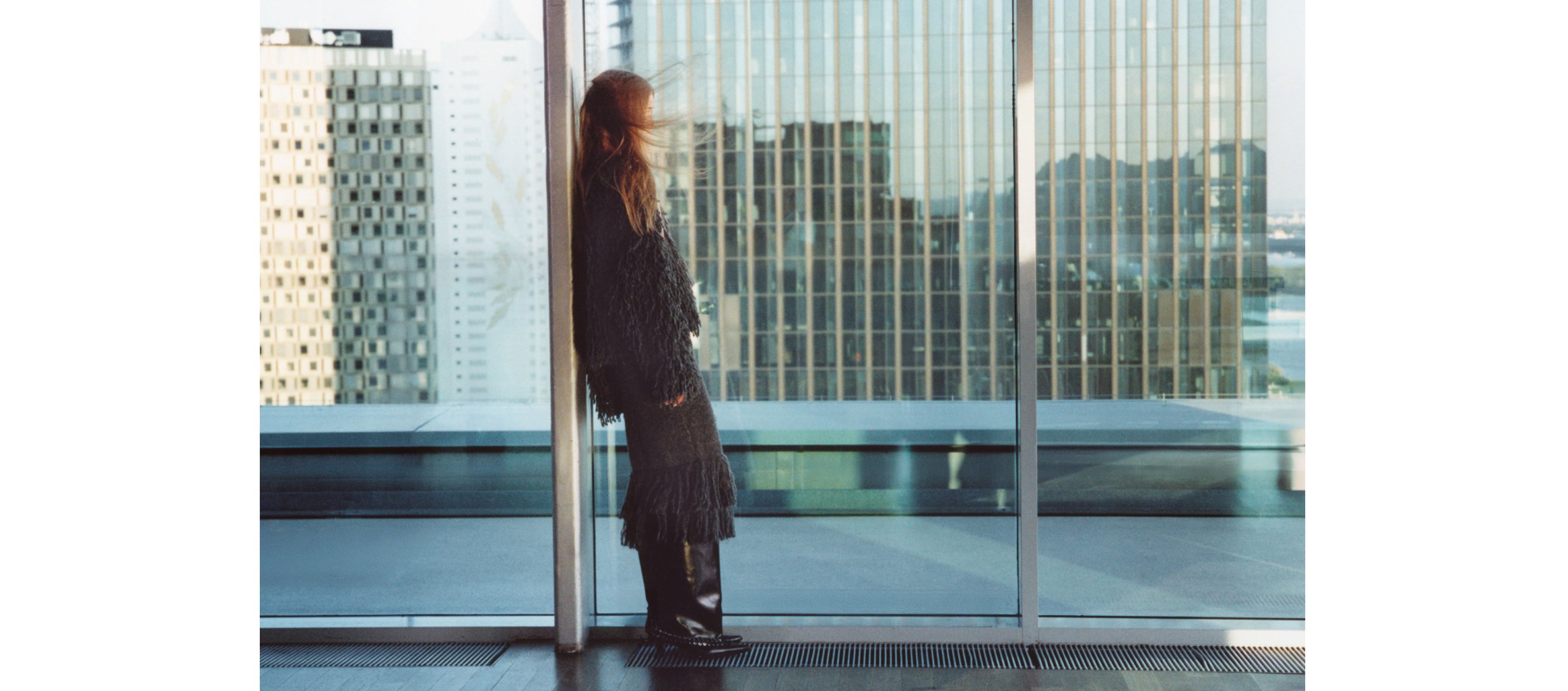 Person in a dark outfit leans against a glass wall, overlooking a cityscape with tall buildings.