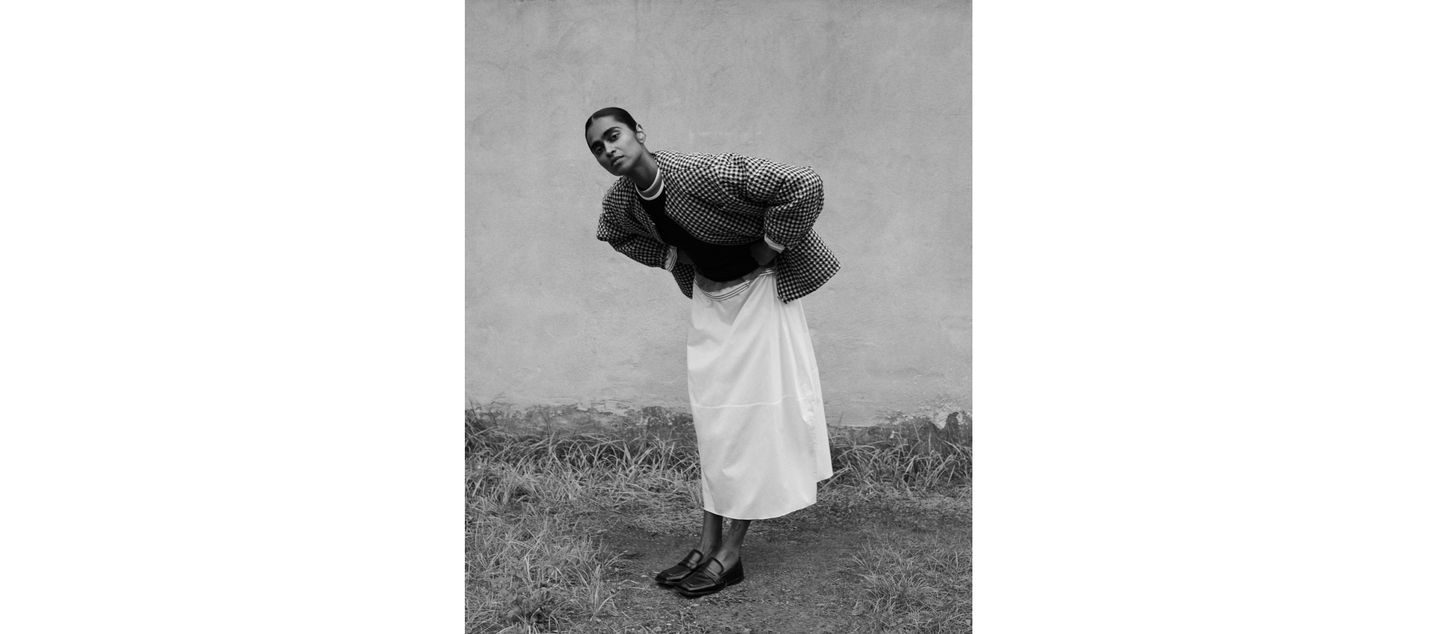 Person in a patterned jacket and long skirt leans forward against a plain outdoor wall, standing on grass, wearing loafers.