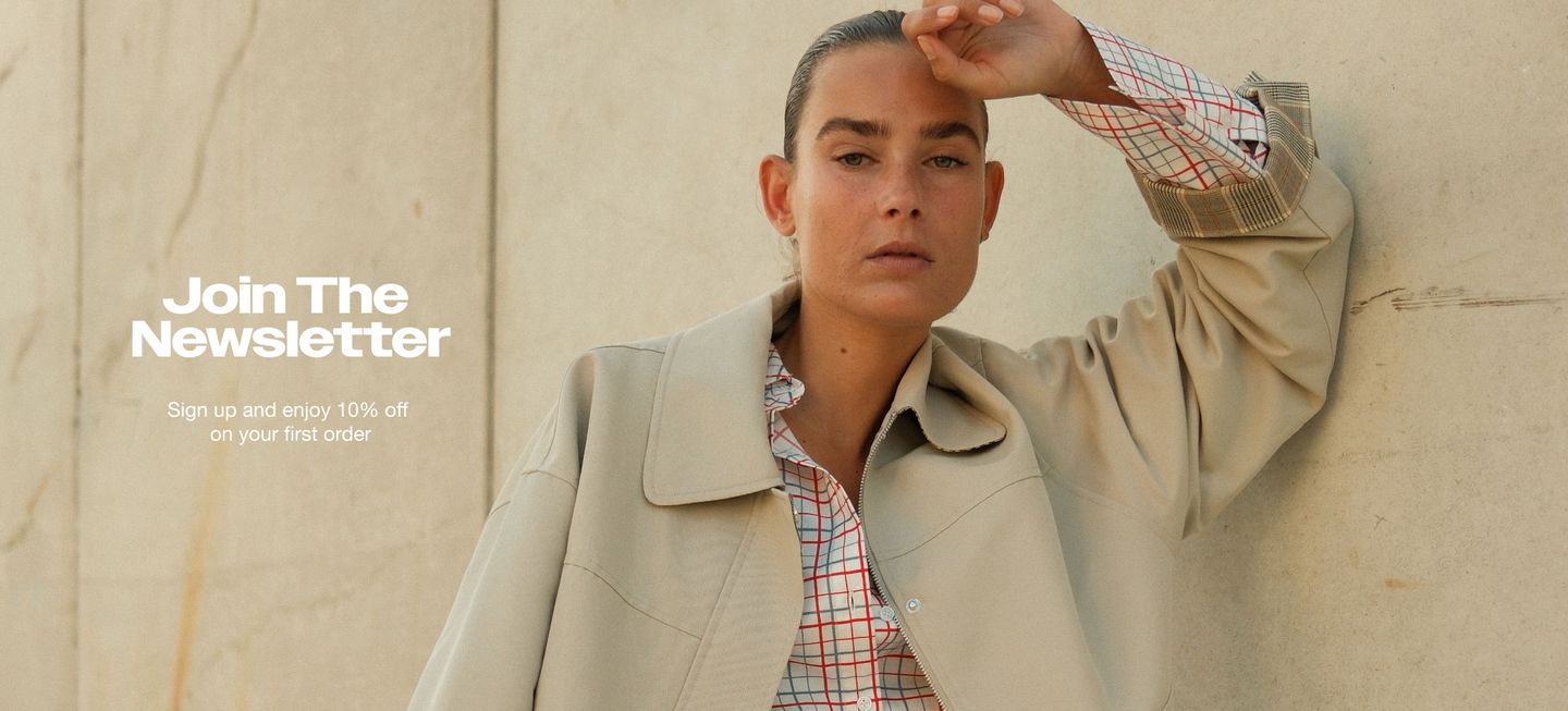 Person in beige coat and checkered shirt leans against a wall. Text reads, "Join The Newsletter" and "Sign up and enjoy 10% off your first order."