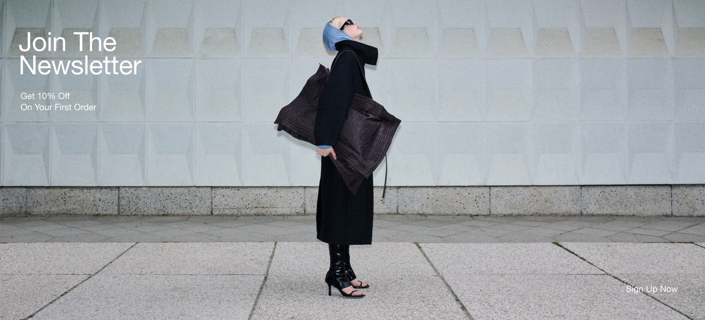 A person in a long black coat and blue headscarf with a large bag poses against a textured wall. Text reads "Join The Newsletter – Get 10% Off."
