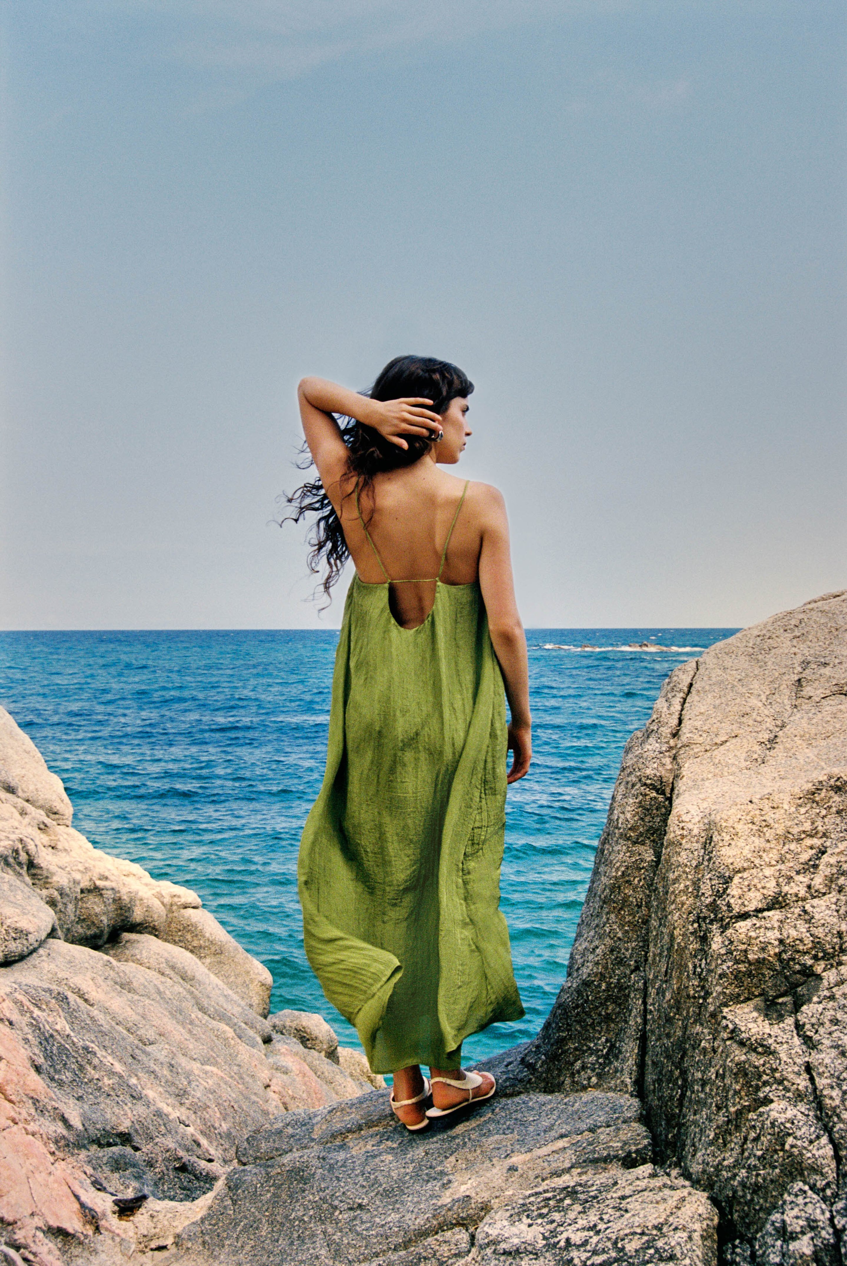 A woman in a flowing green dress stands on rocky cliffs, gazing at the ocean under a clear blue sky.