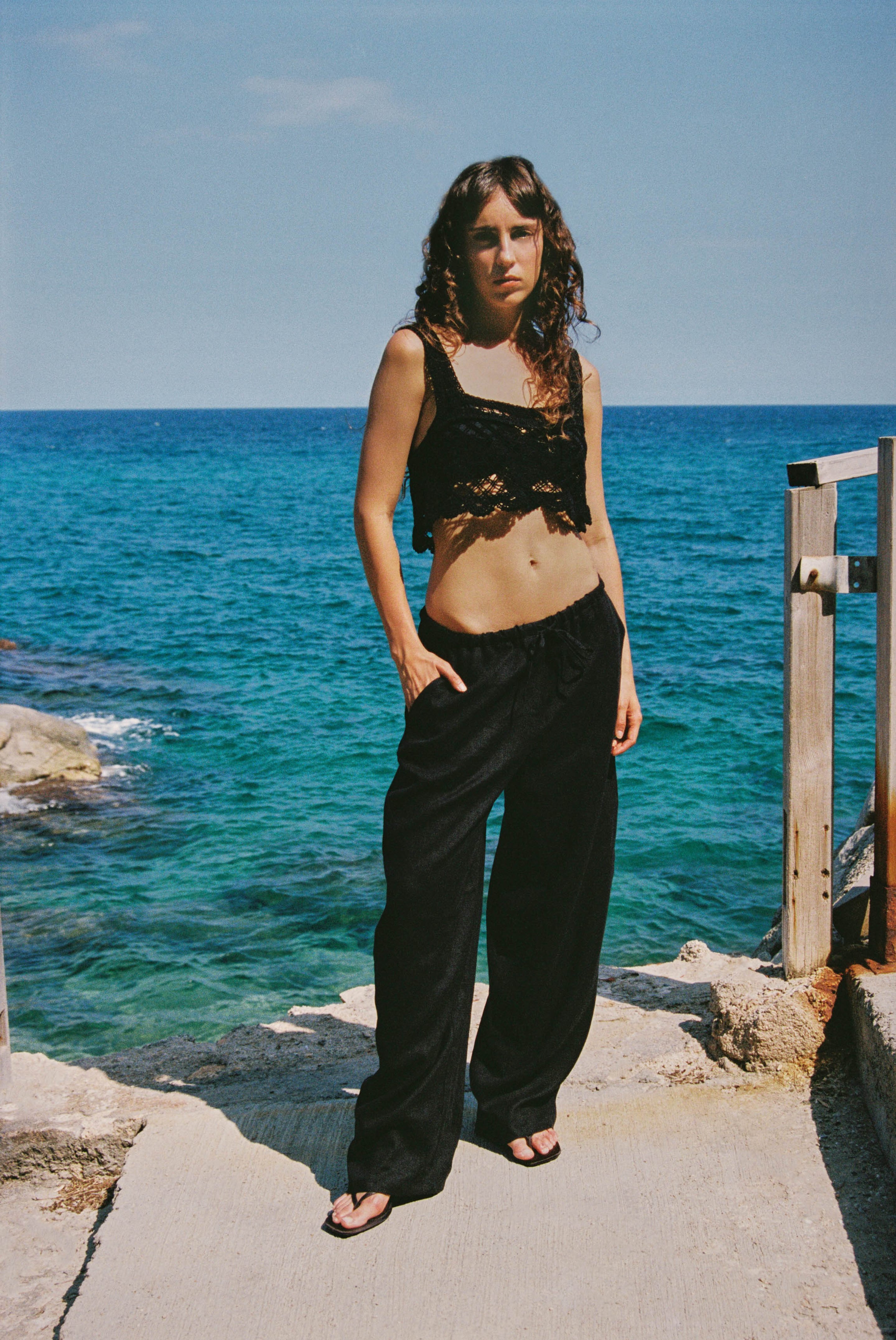 Frau am Strand mit schwarzem, gehäkeltem Crop-Top und schwarzer Hose mit Tunnelzug, im Hintergrund türkisfarbenes Meer
