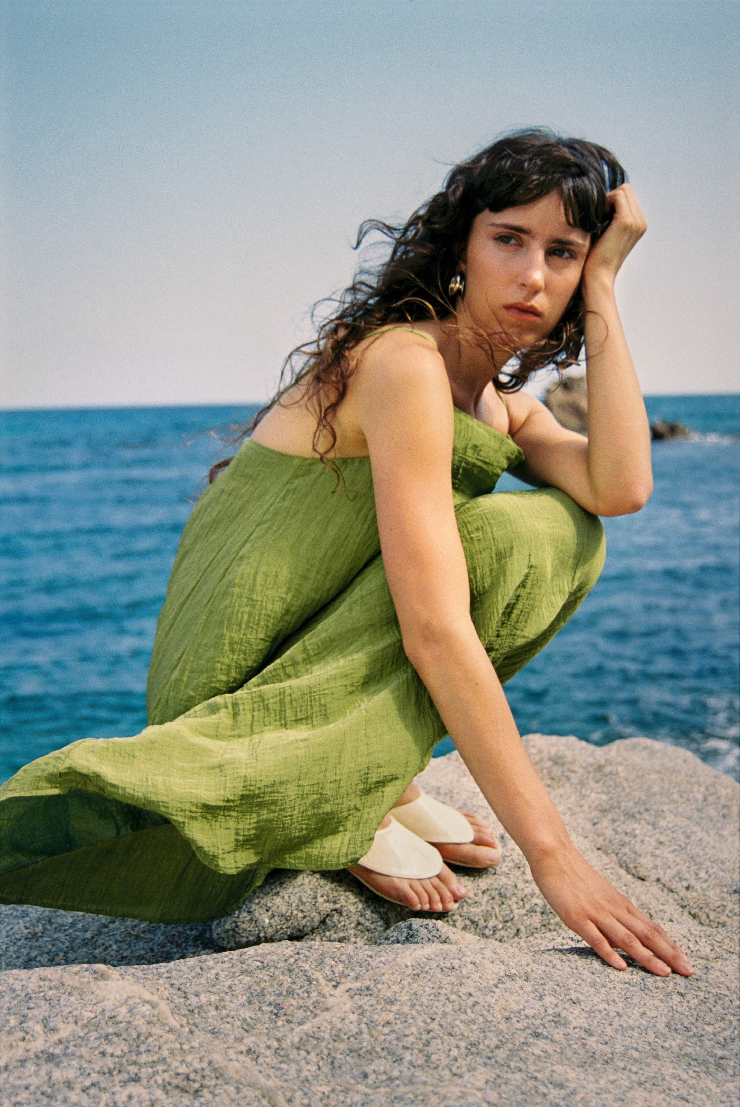 Woman in a green dress sits on a rock by the sea, with long hair flowing in the breeze and a contemplative expression.