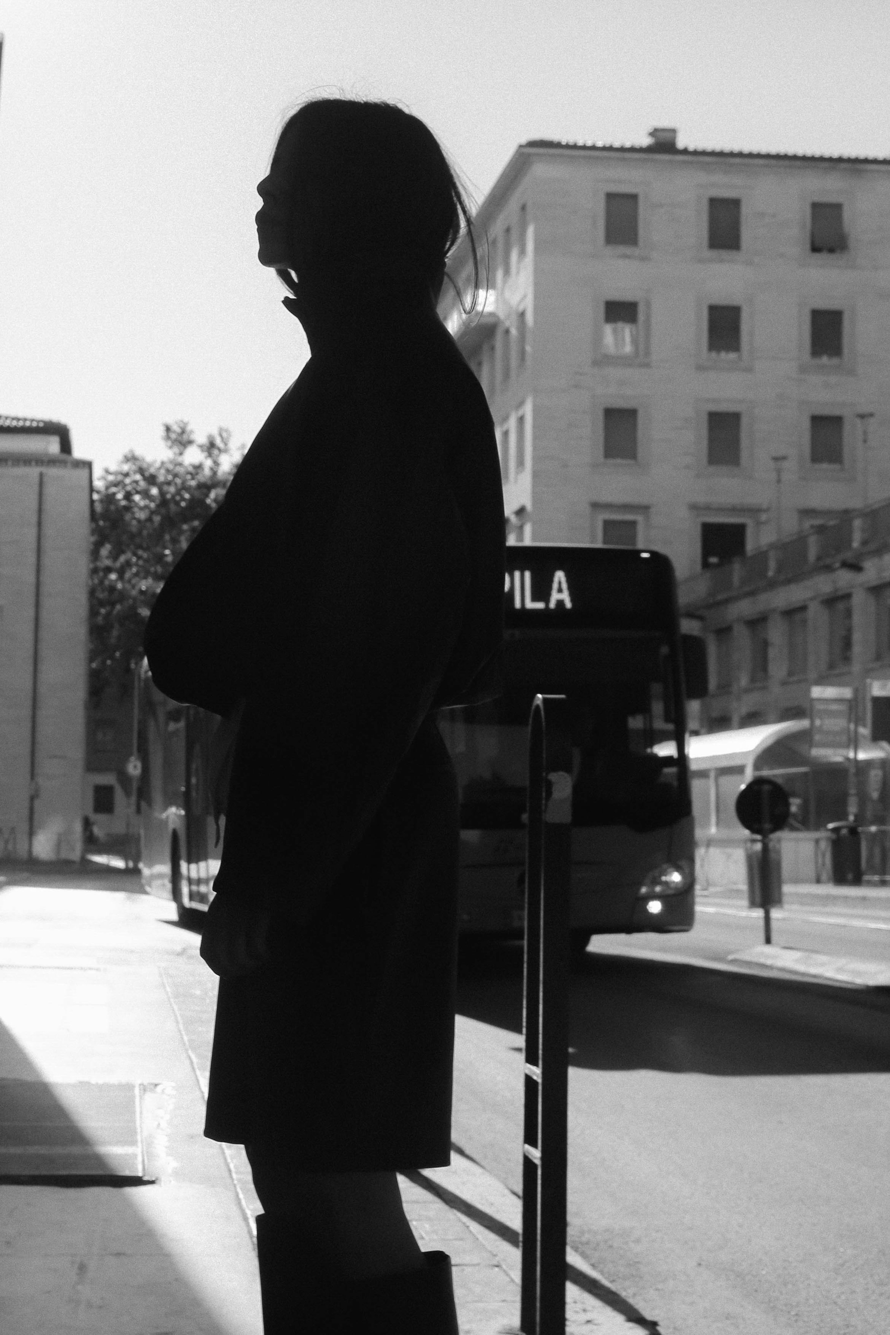 Silhouette of a person in a coat standing near a bus stop, with a bus in the background on a city street. Black and white.
