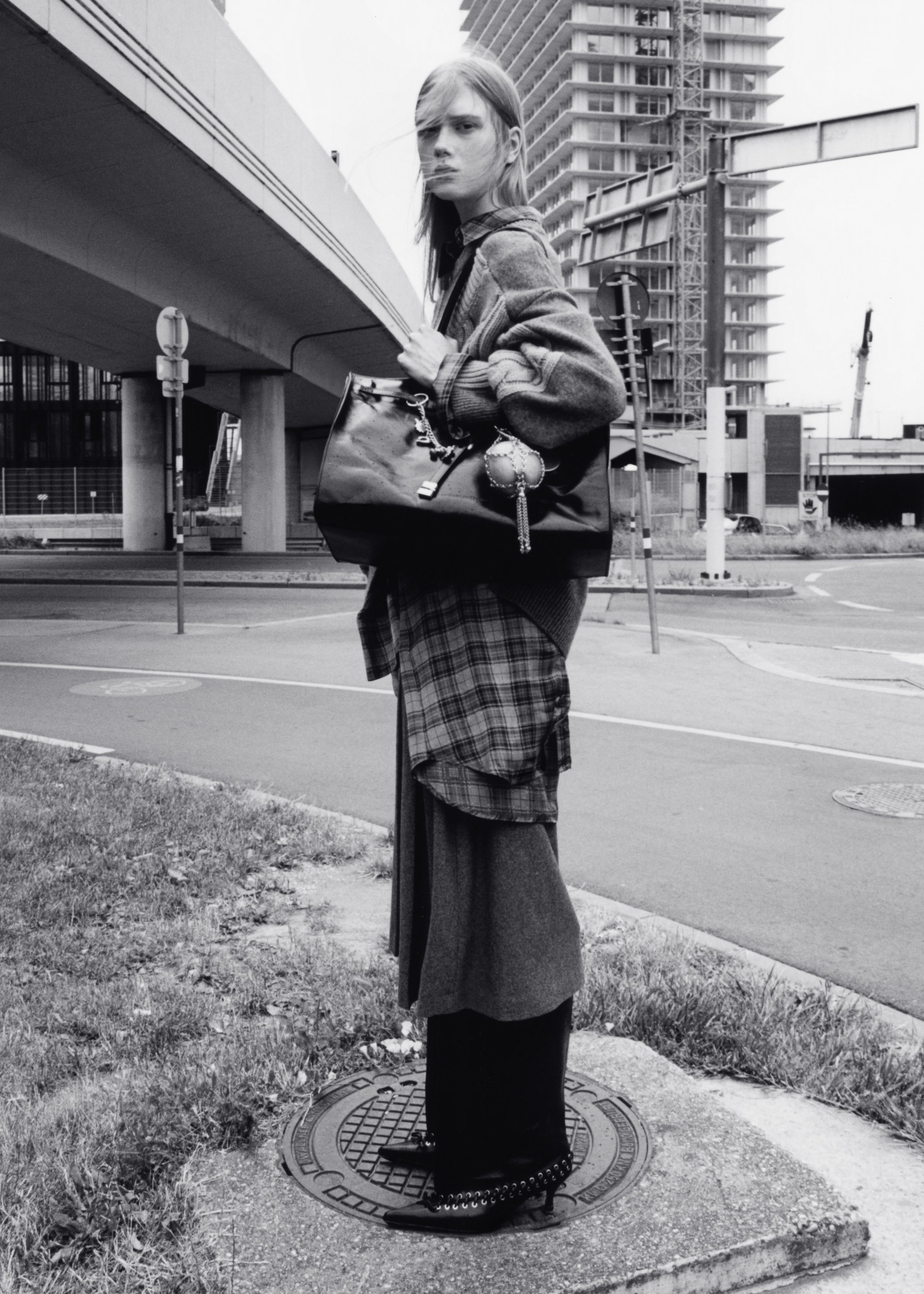 A person stands on a manhole cover in an urban setting, wearing layered clothing and holding a large handbag, with buildings in the background.