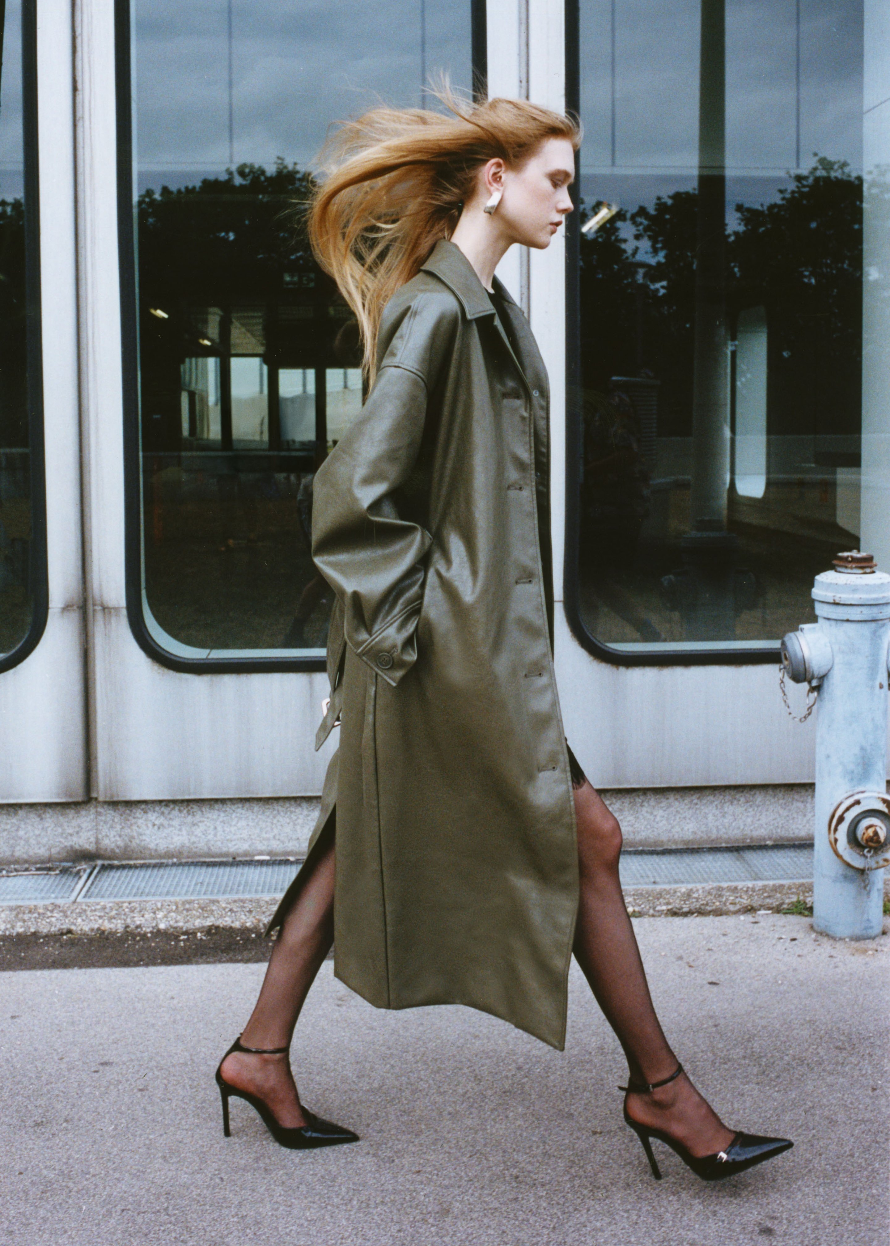A woman with long hair walks confidently in a green trench coat and black heels, passing a building with large windows.