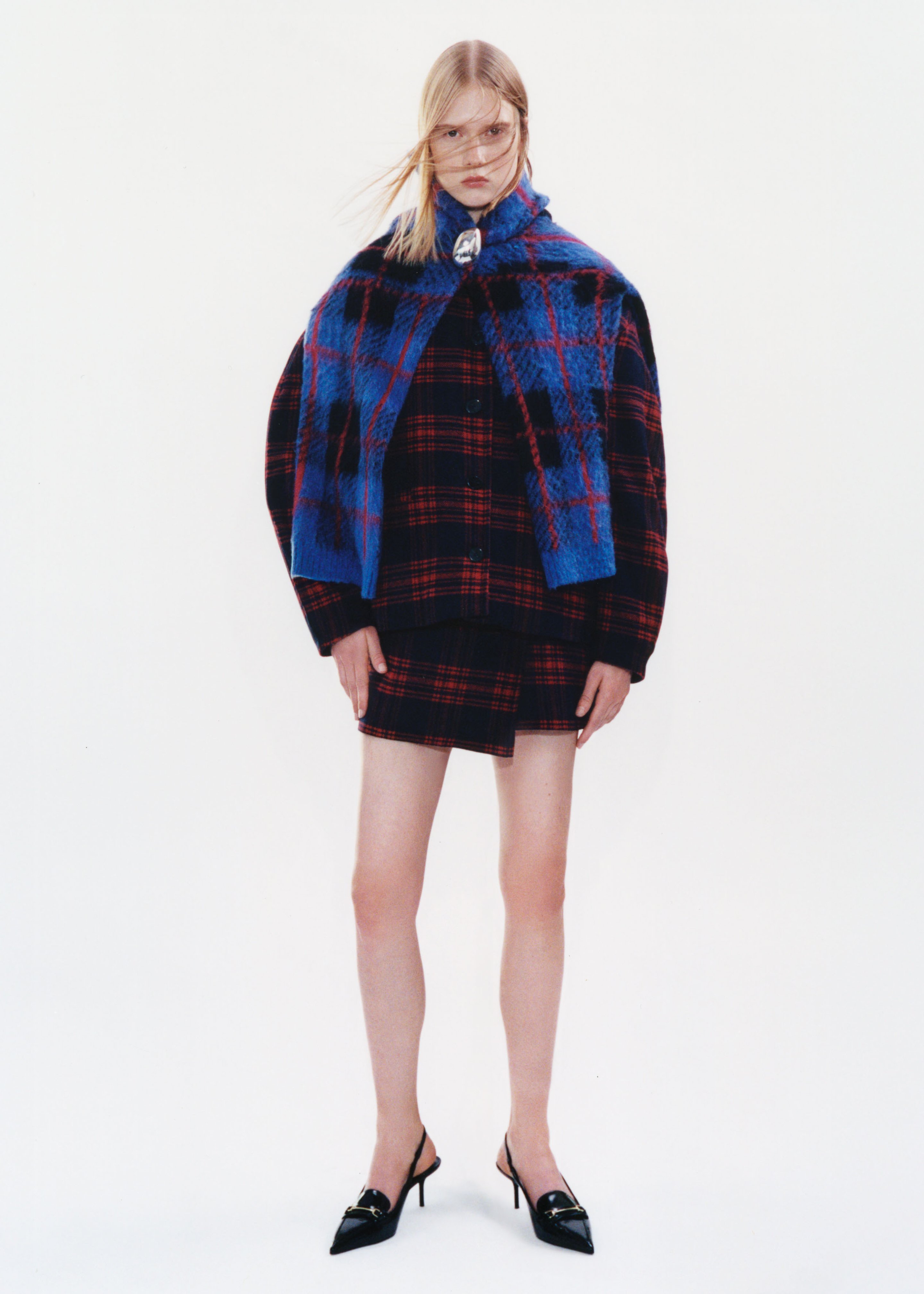 Model in plaid jacket and skirt, with a blue shawl, wearing black pointed heels, stands against a plain white background.