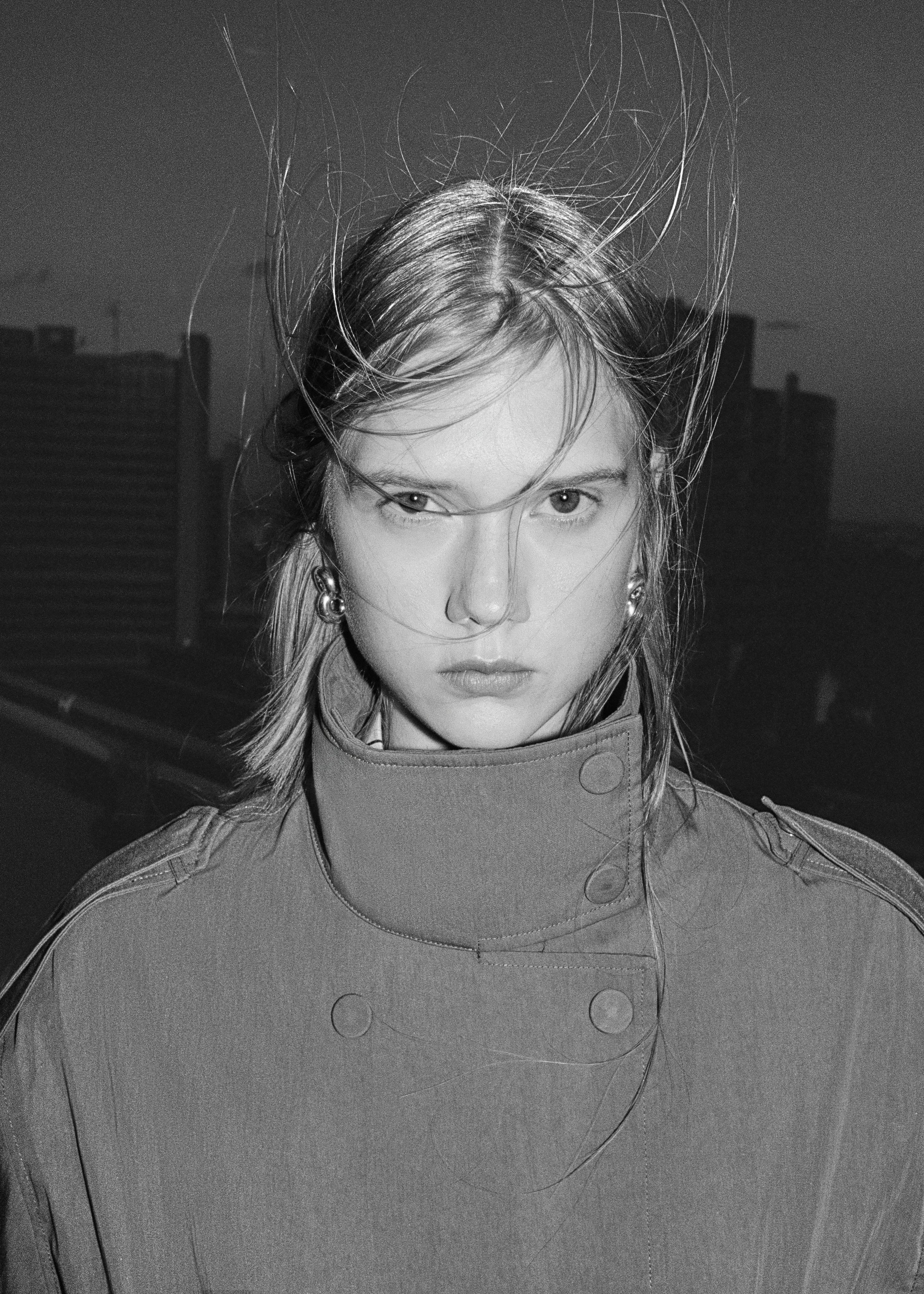 Black and white photo of a person with windblown hair, wearing a high-collared coat, standing against a cityscape background.