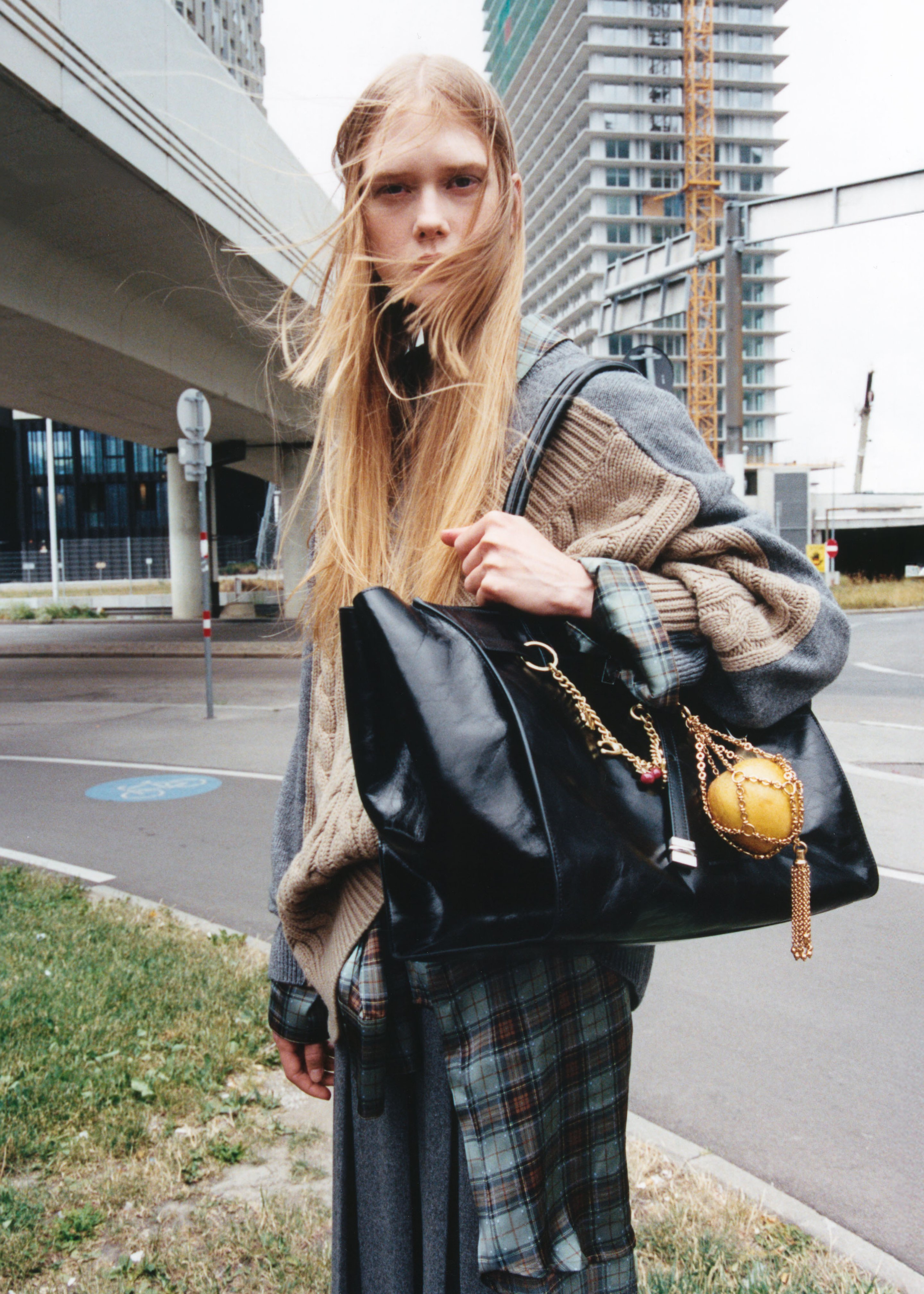 Person with long hair in layered clothing holds a large black bag, standing outdoors near a road and modern buildings.