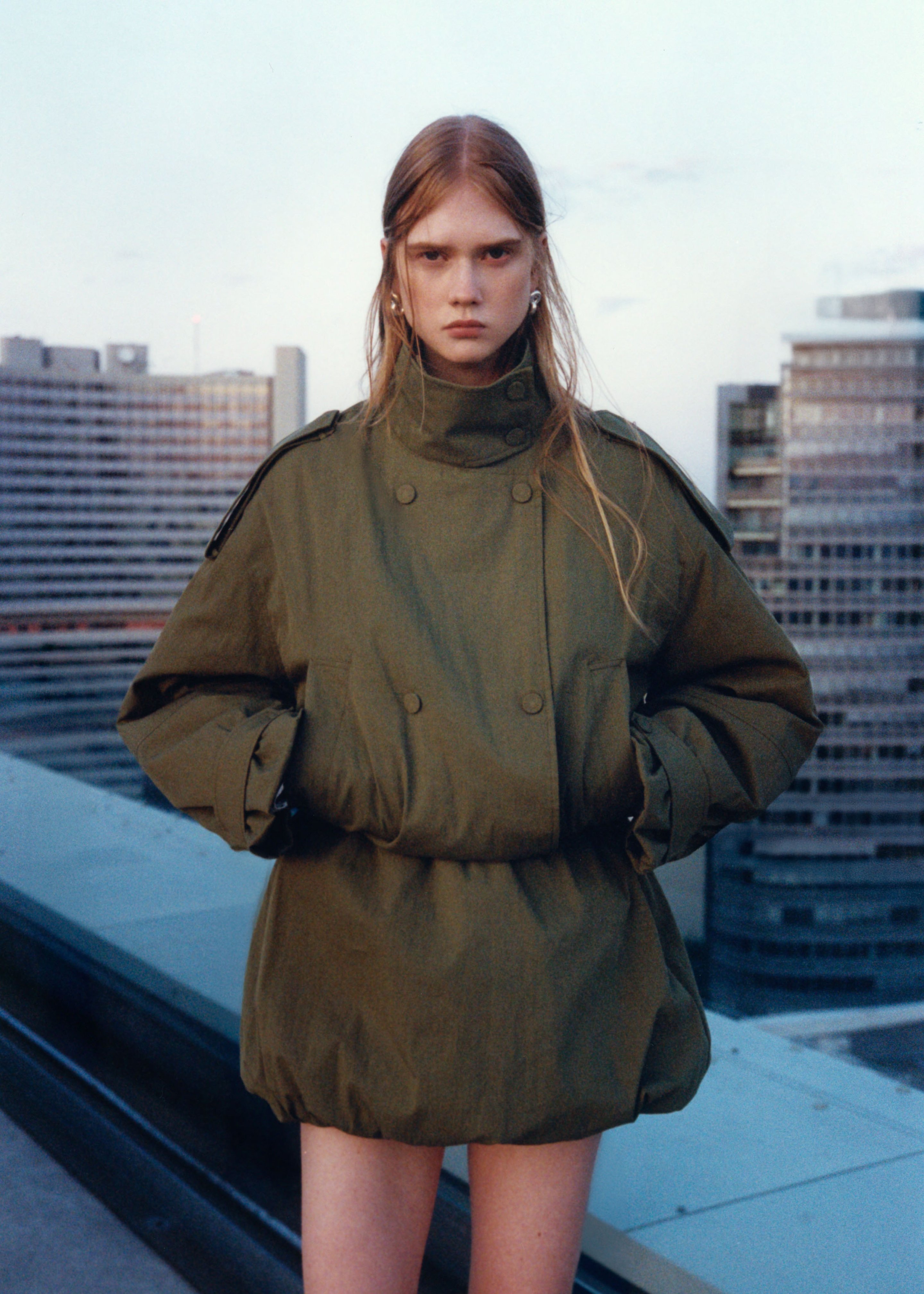 Person in oversized green coat stands on a rooftop with a cityscape backdrop at dusk.