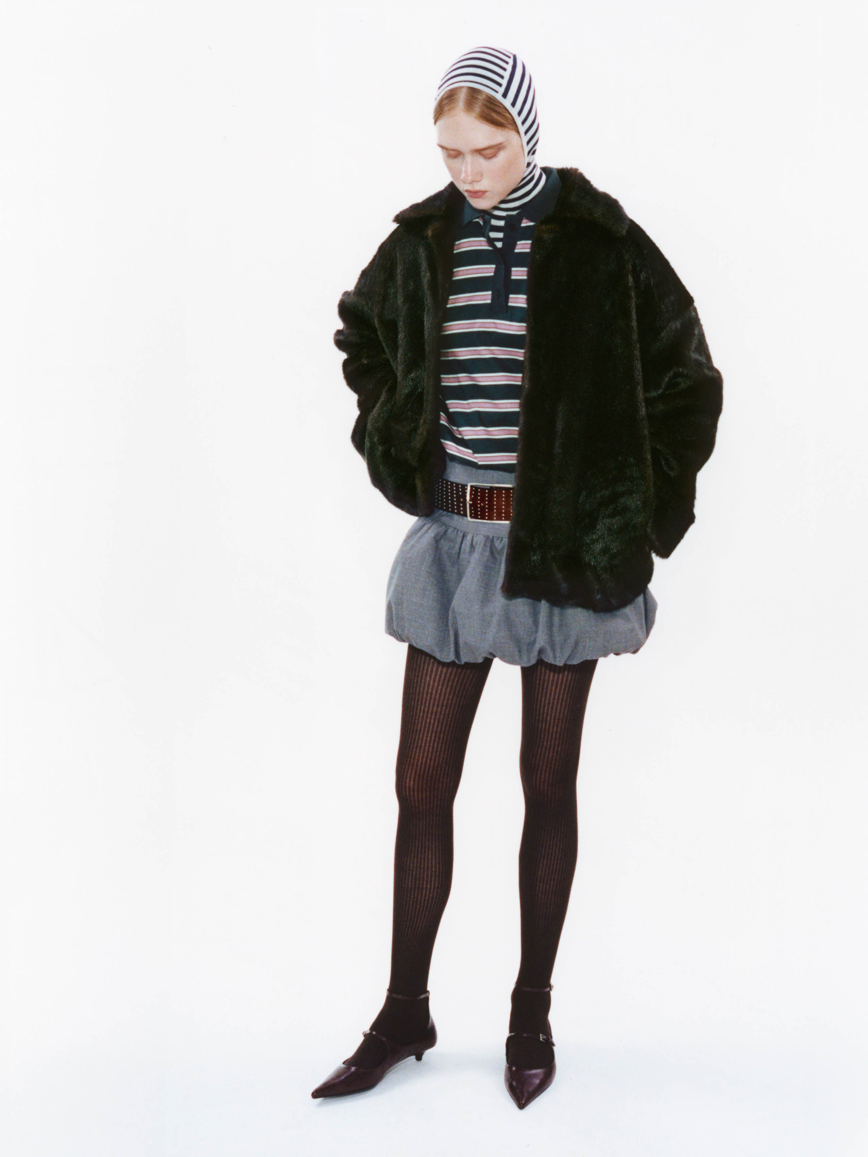 A person in a striped top, black jacket, and skirt stands with hands in pockets, wearing a headscarf, tights, and pointed shoes.