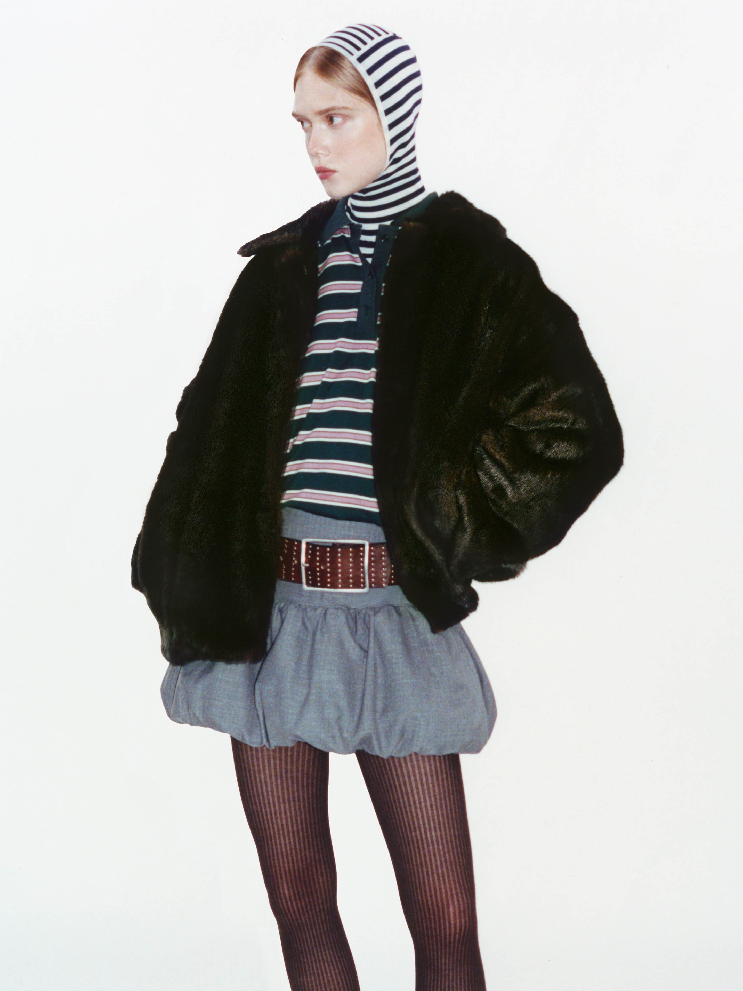 Person in a fur jacket, striped shirt, and skirt with striped tights and a striped hood, posing against a plain background.