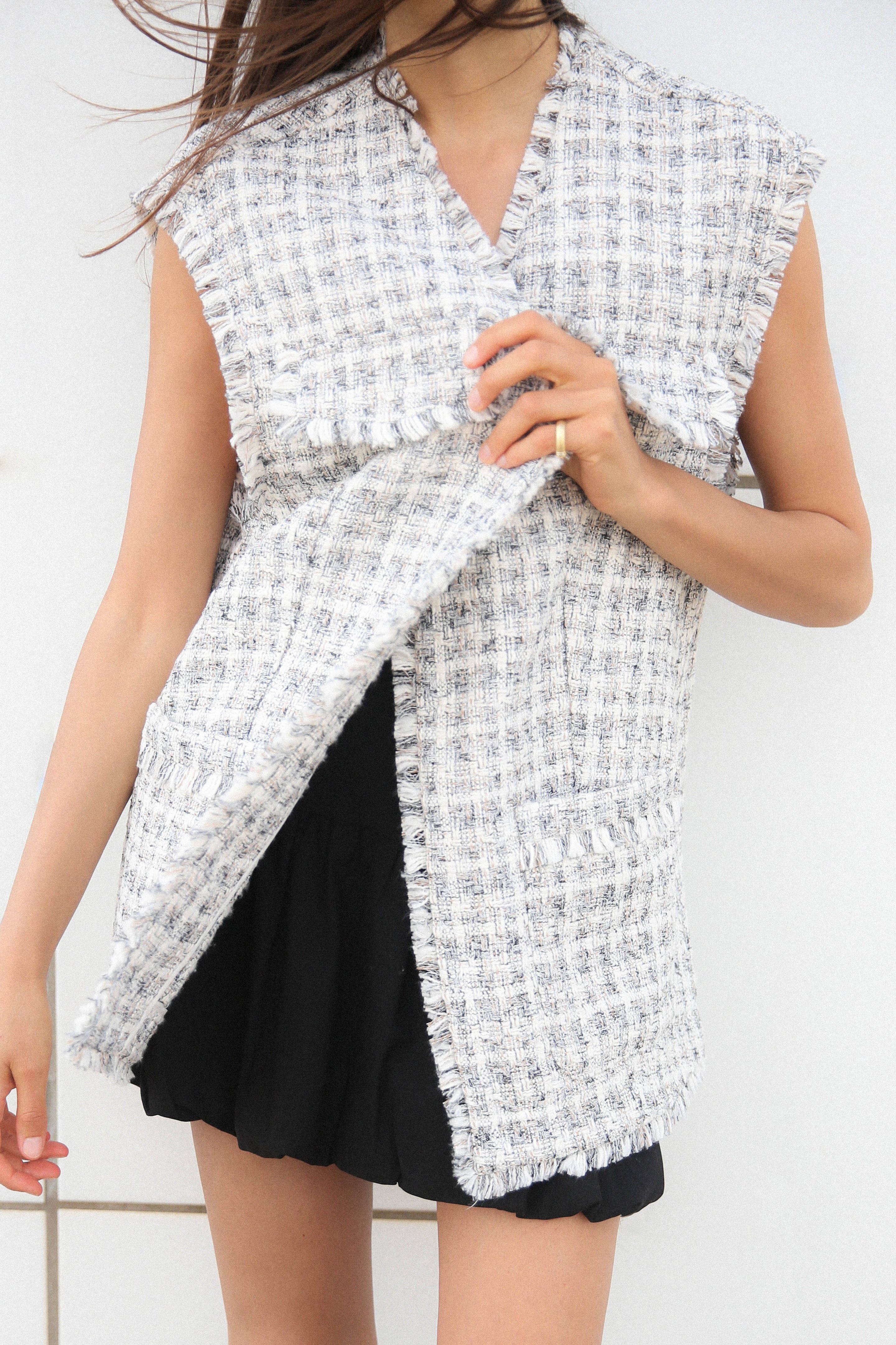 Woman wearing a sleeveless, textured tweed vest over a black skirt, with wind blowing her hair and hand adjusting the vest.