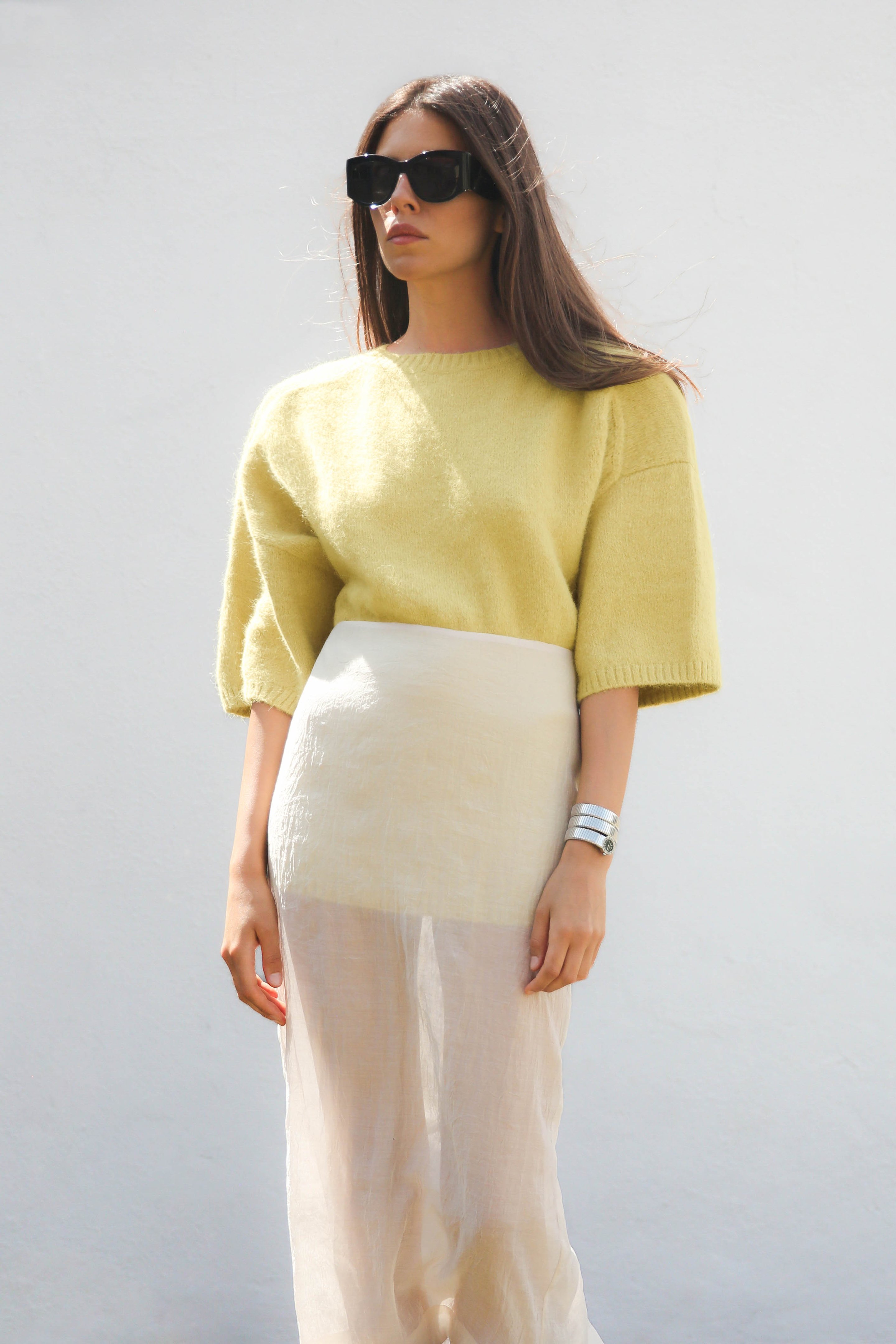 Woman in sunglasses wearing a yellow sweater and sheer white skirt, standing against a plain white background.