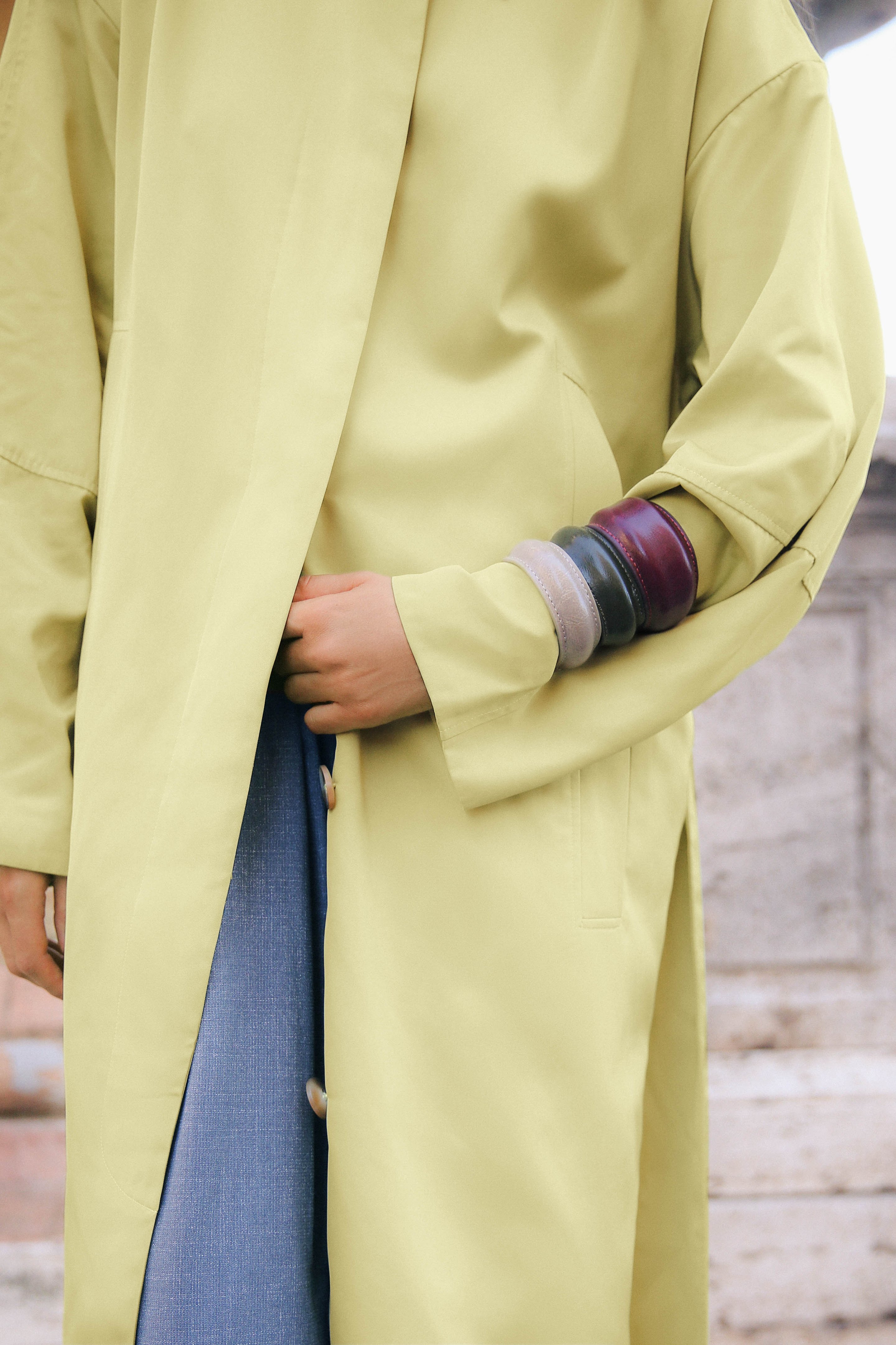 Person in a light green coat and blue jeans, wearing multiple colorful bangles on one arm, standing outdoors.