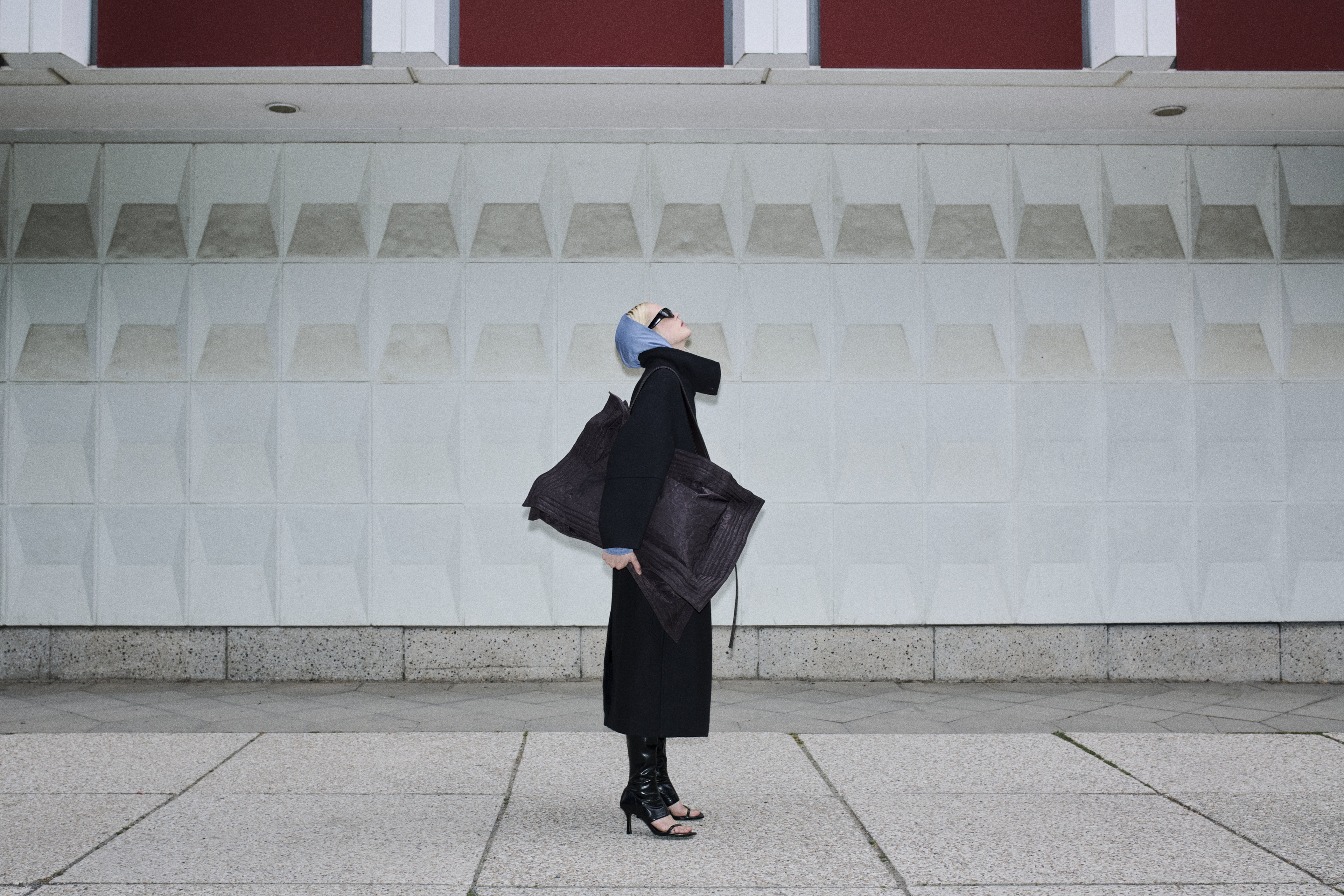 A person in a stylish outfit, wearing a headscarf and sunglasses, stands against a geometric wall, carrying a large textured bag.
