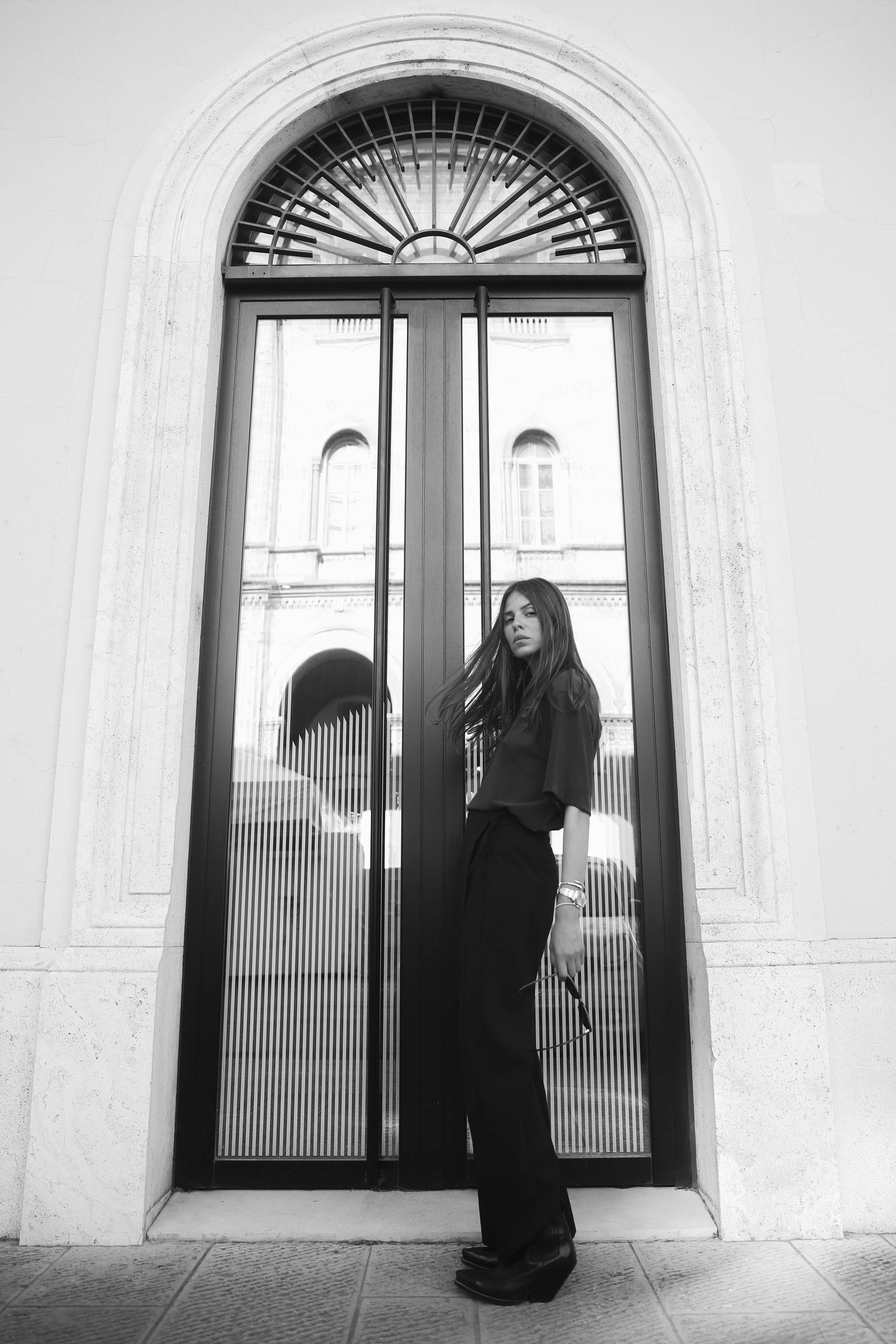 Woman standing in front of an arched glass door, wearing dark clothing, with a building reflection visible in the door. Black and white image.