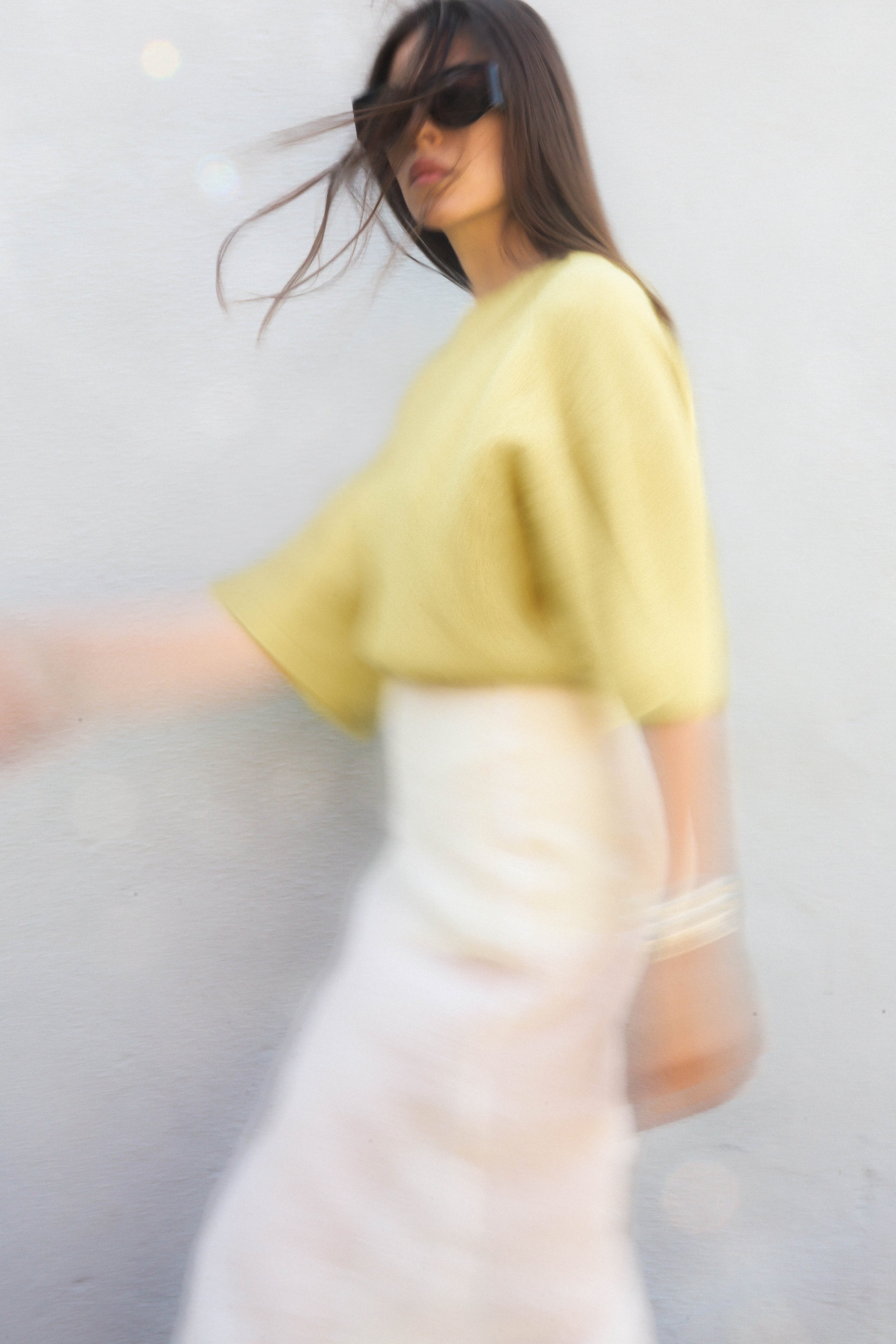 Blurred image of a woman in a yellow top and white skirt, wearing sunglasses, walking against a plain background.