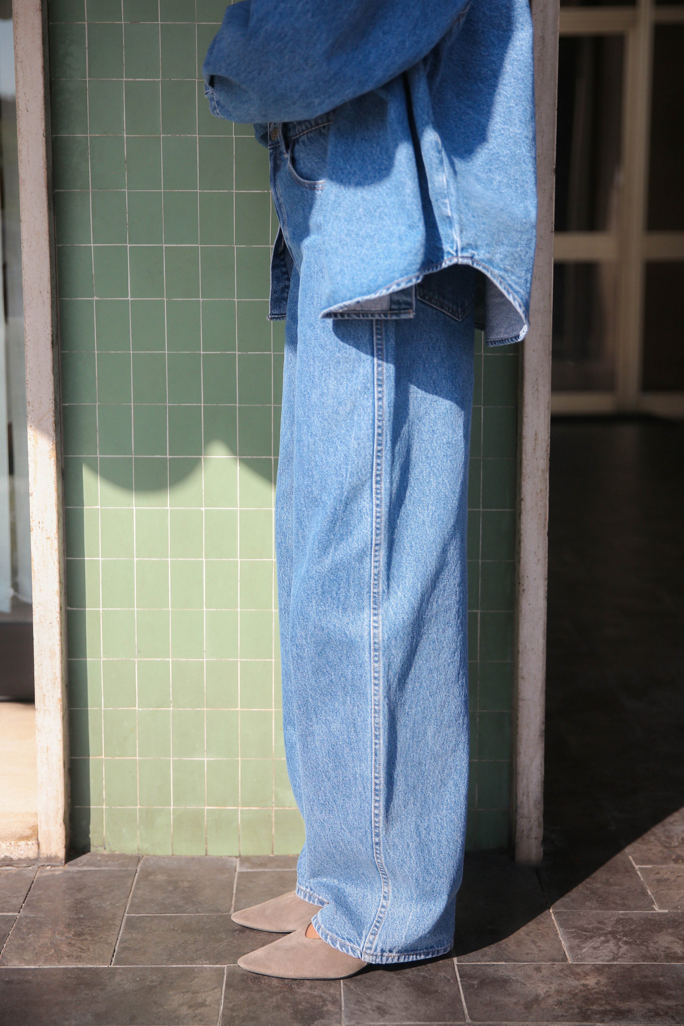 Person in blue denim jacket and wide-leg jeans stands on a dark floor, next to a green tiled wall, wearing pointed shoes.