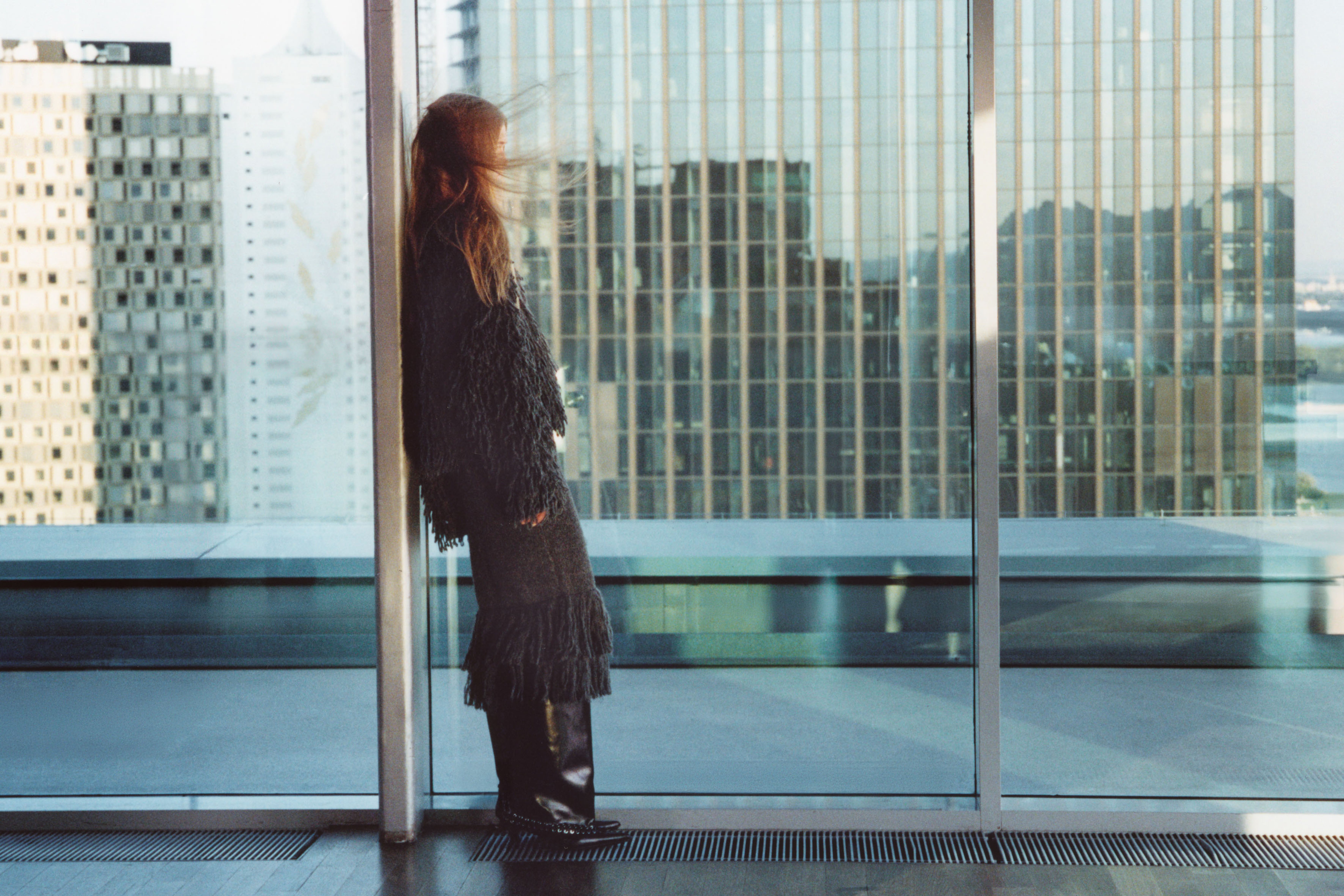 Person in a fringed outfit leans against a glass wall on a balcony, with city skyscrapers visible in the background.