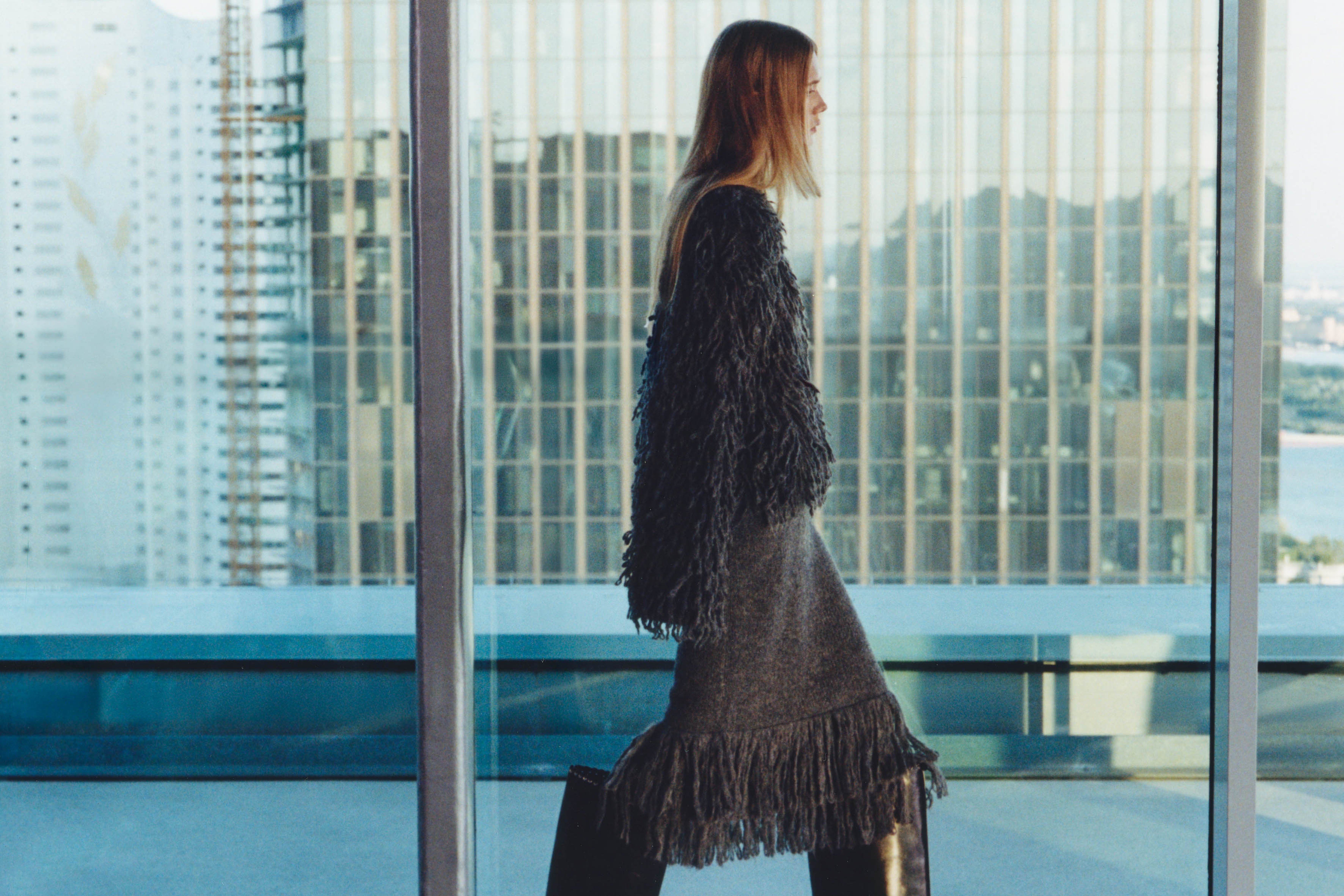 A person in a fringed outfit stands by a large window, with city skyscrapers reflecting sunlight in the background.
