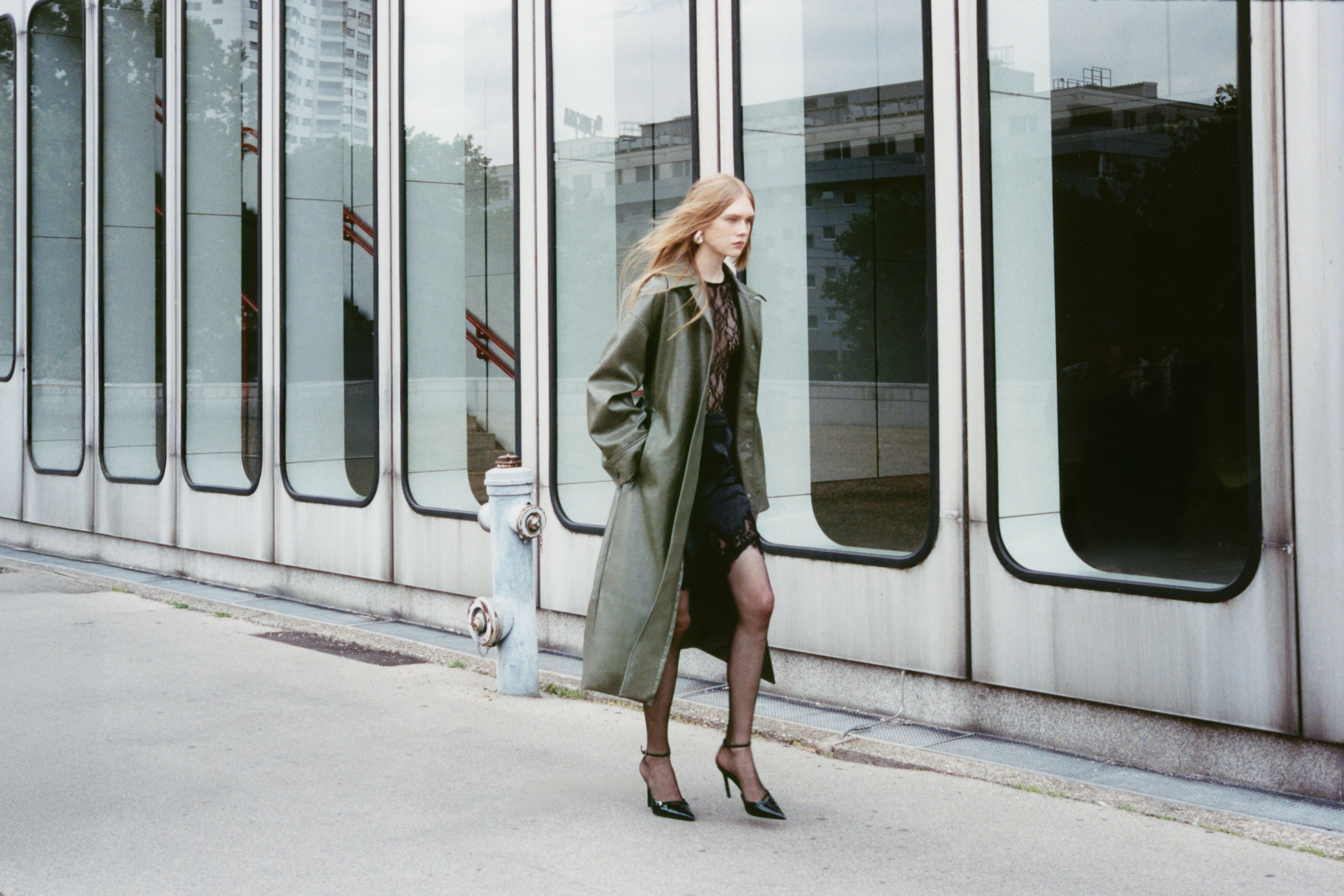 A person in a long green coat and heels walks confidently on a city sidewalk beside modern glass windows.