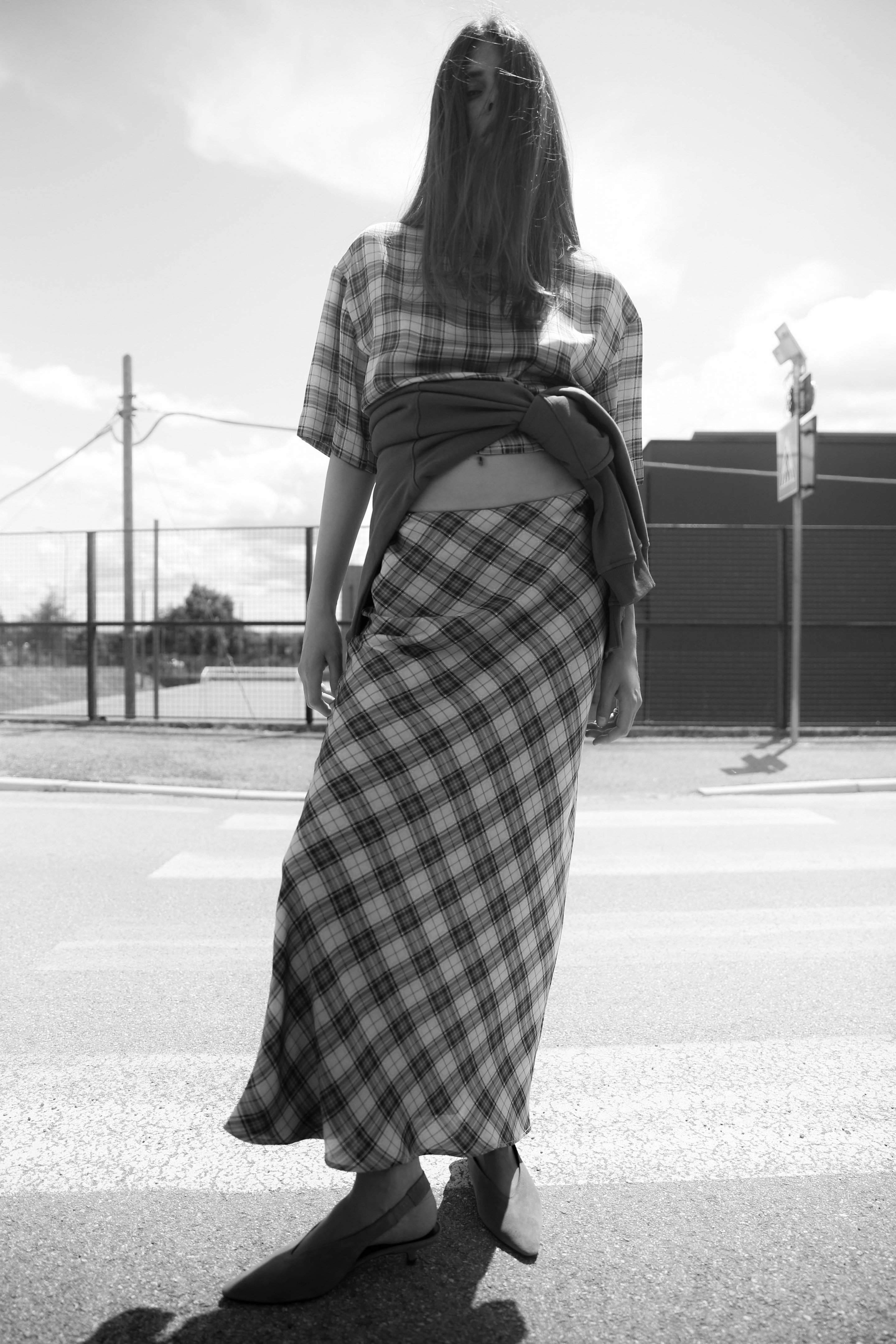 Woman in plaid outfit stands on a street, hair covering face, with a building and fence in the background. Black and white photo.
