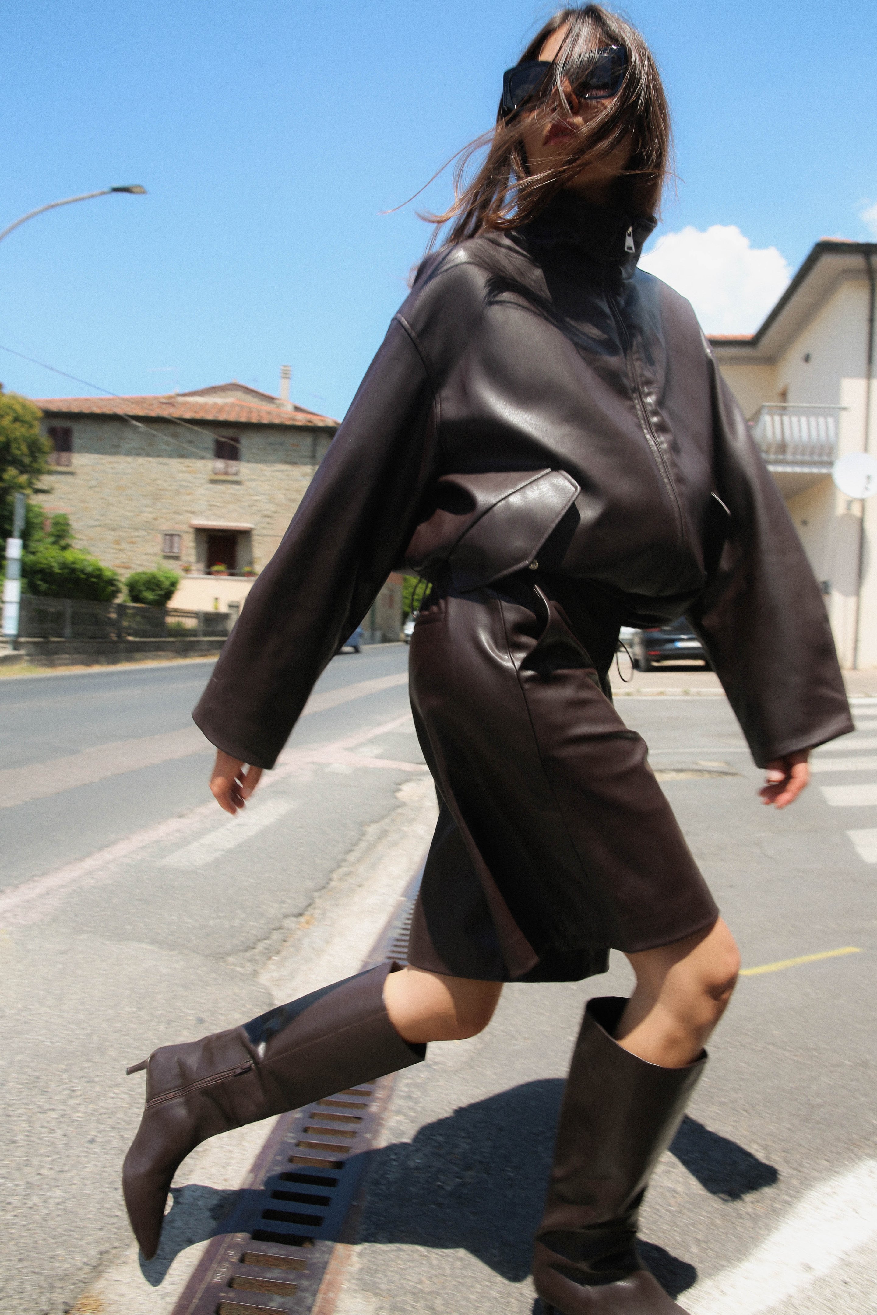 Woman in oversized dark leather jacket, skirt, and boots walking on a sunny street, with sunglasses and hair flowing.