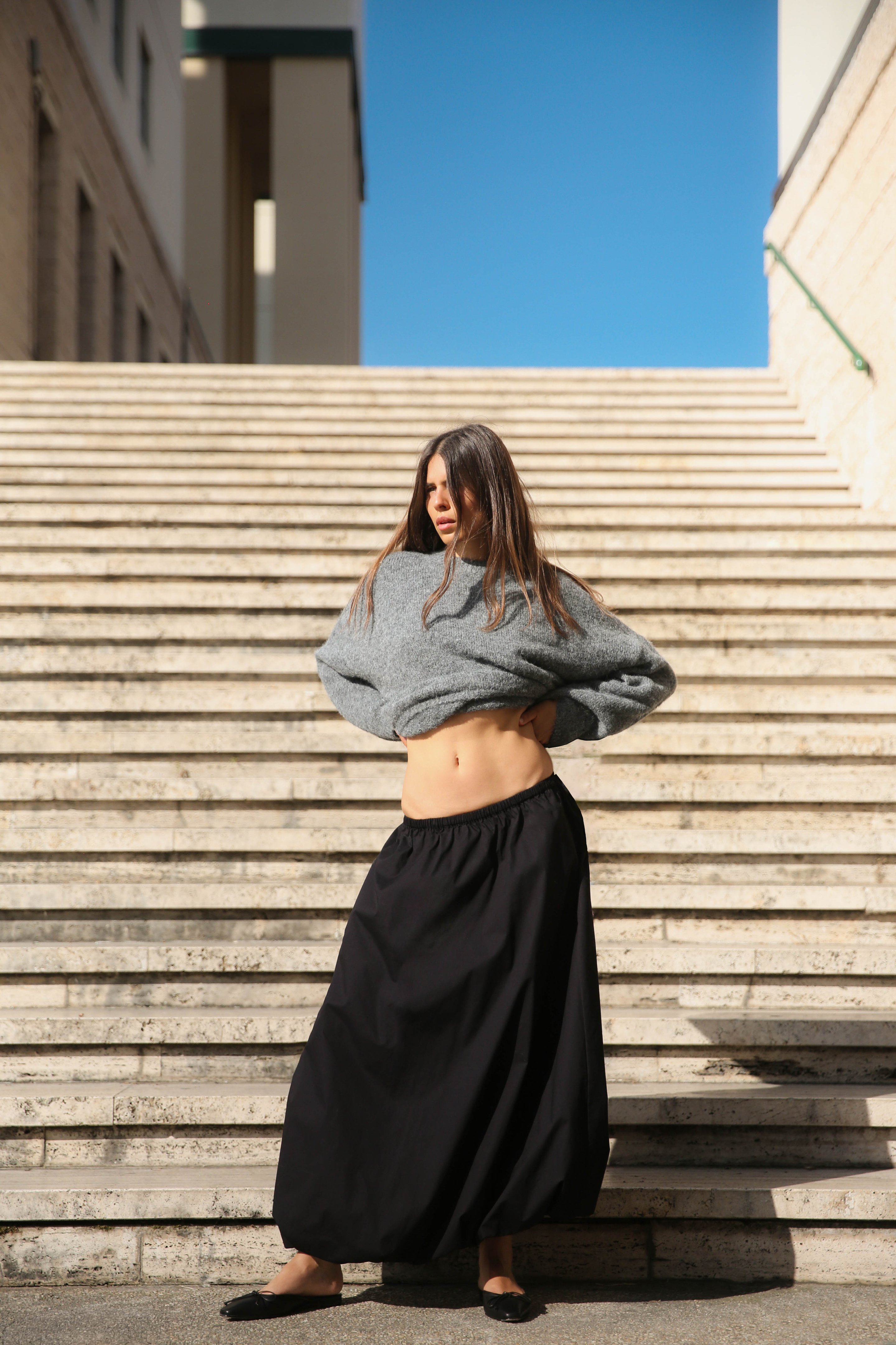A woman stands on stone steps, wearing a gray sweater and long black skirt, under a clear blue sky.