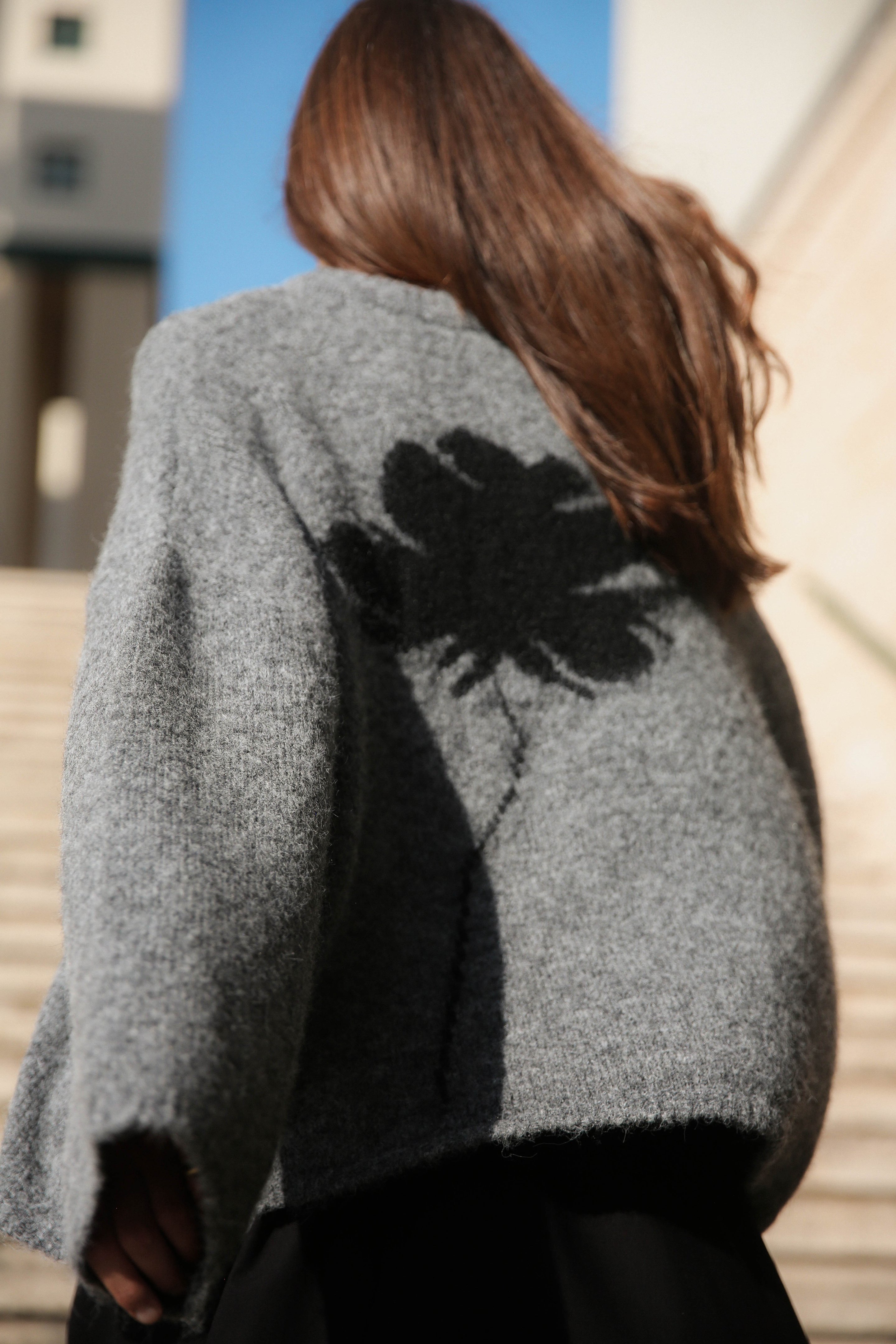 Person with long hair wearing a gray sweater with a black flower design on the back, walking up steps under a clear blue sky.