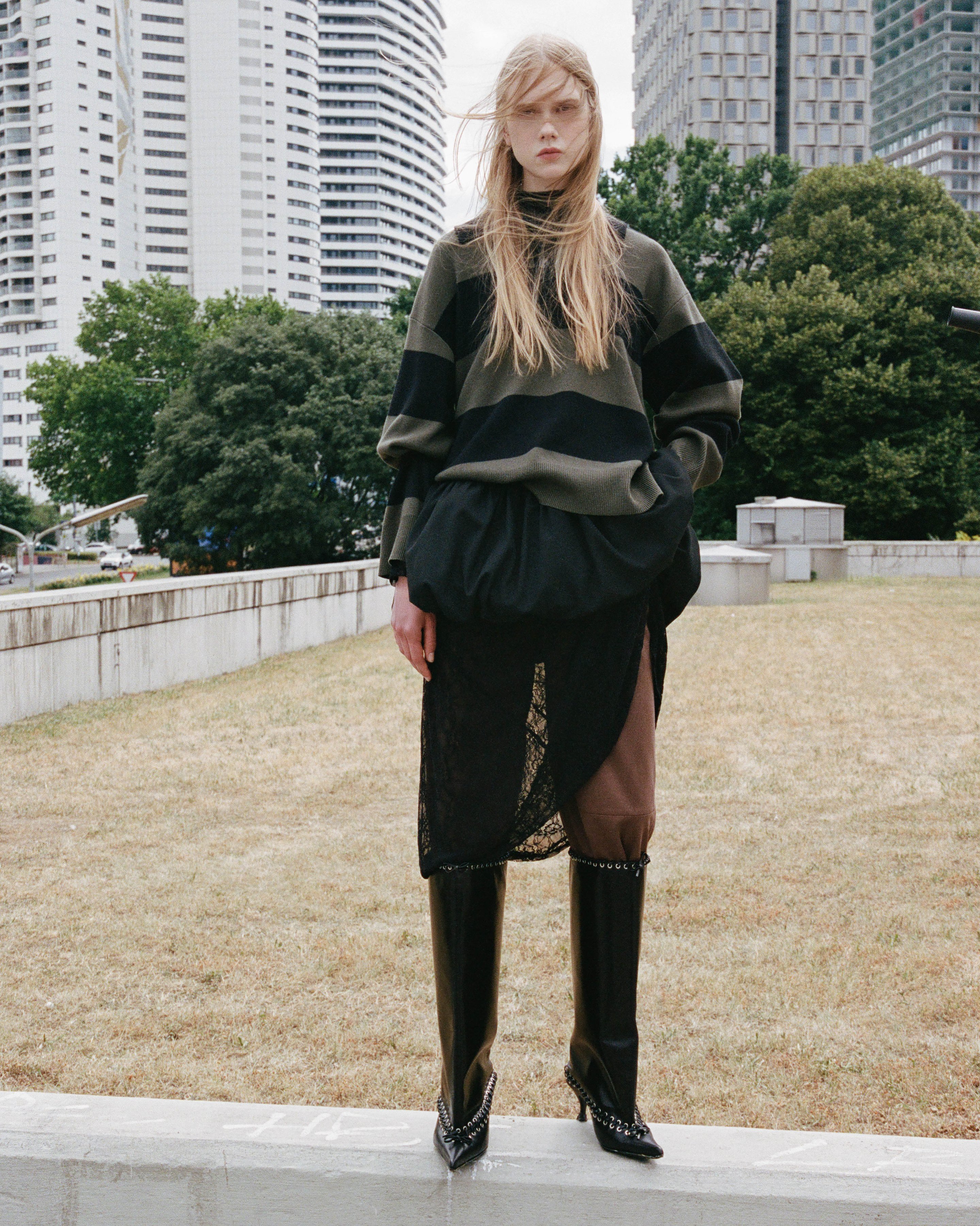 A person with long hair stands outdoors, wearing a striped sweater, layered skirt, and tall boots. Modern buildings and trees are in the background.