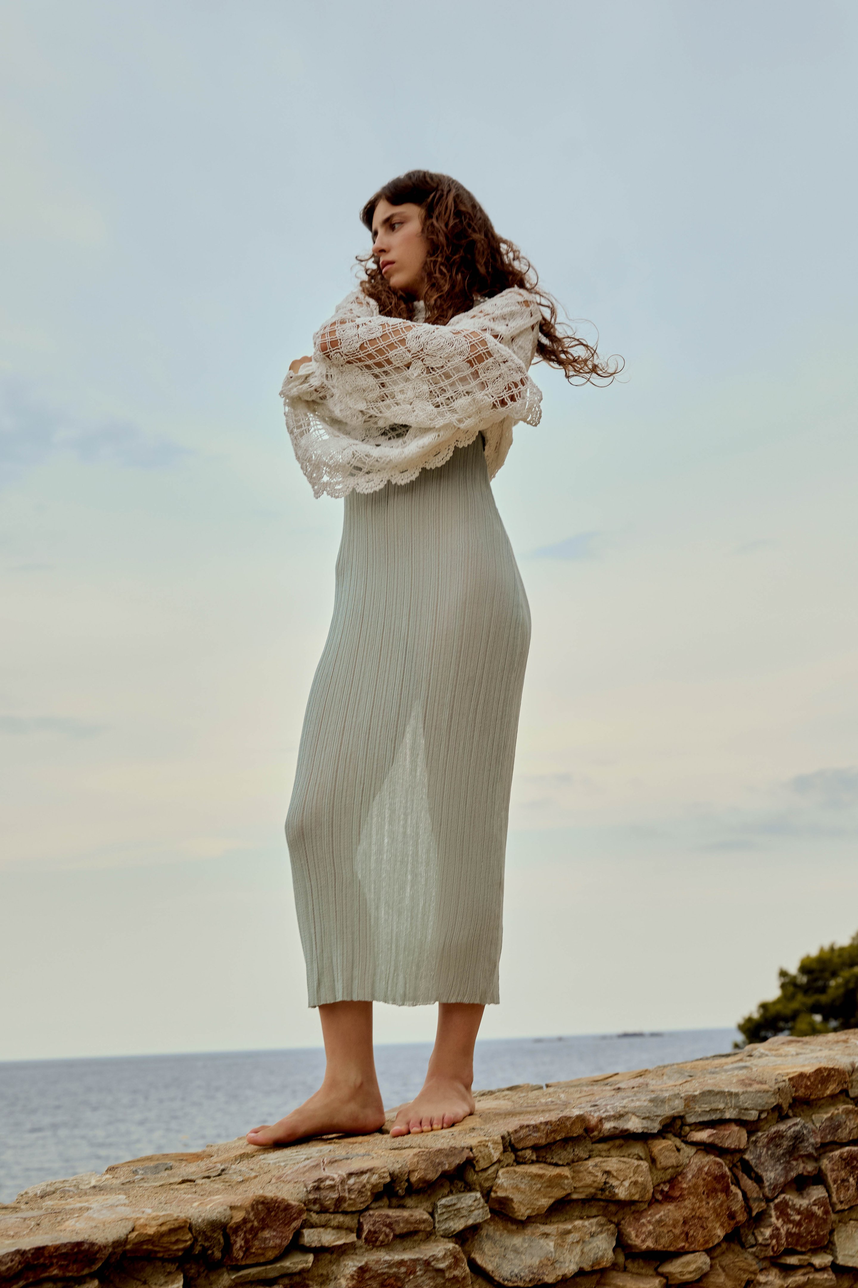 Woman in a long, light dress and lace shawl stands barefoot on a stone wall, gazing at the sea under a cloudy sky.