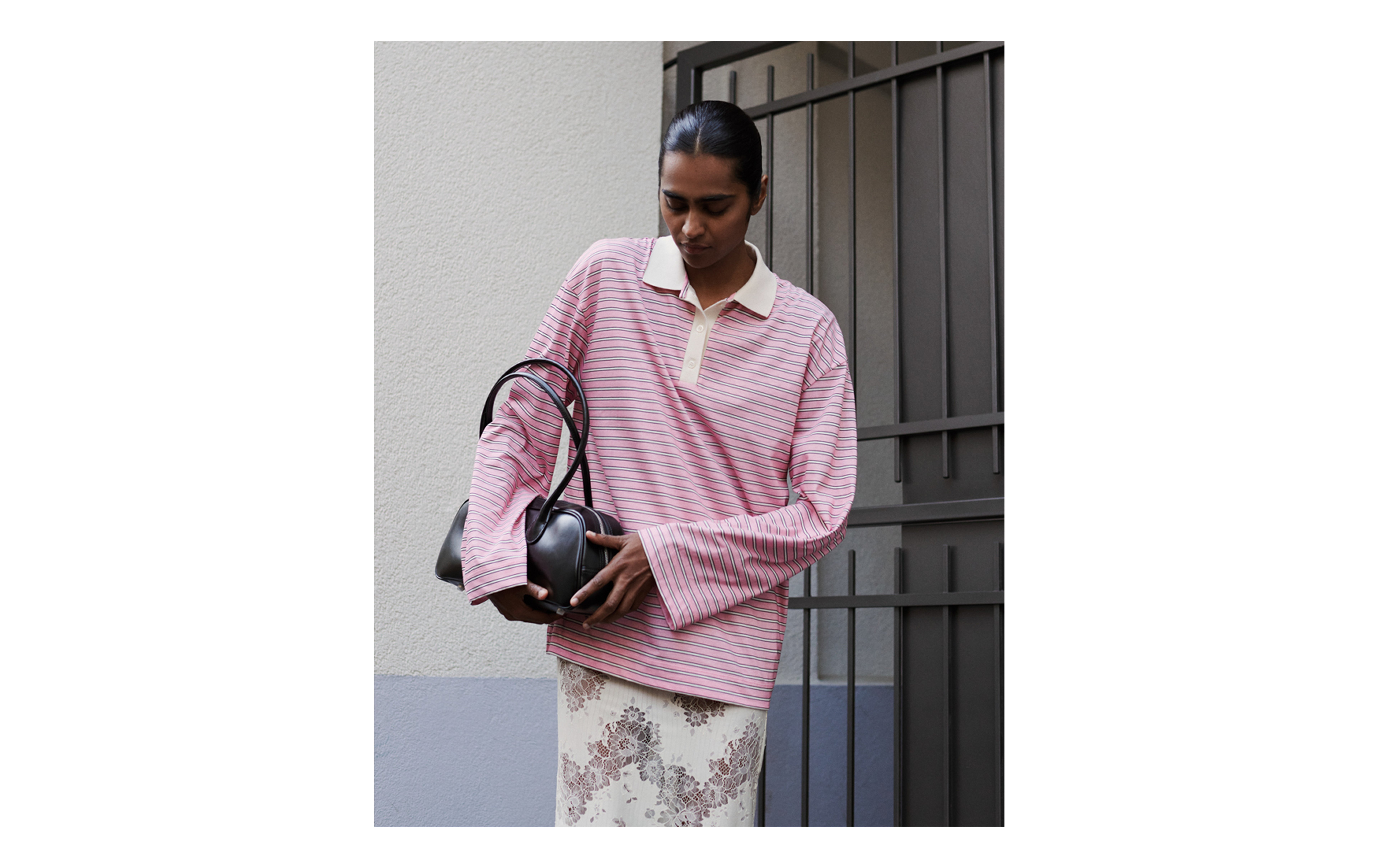 Person in a pink striped shirt and floral skirt stands against a gray wall, holding a black handbag, with a metal gate in the background.
