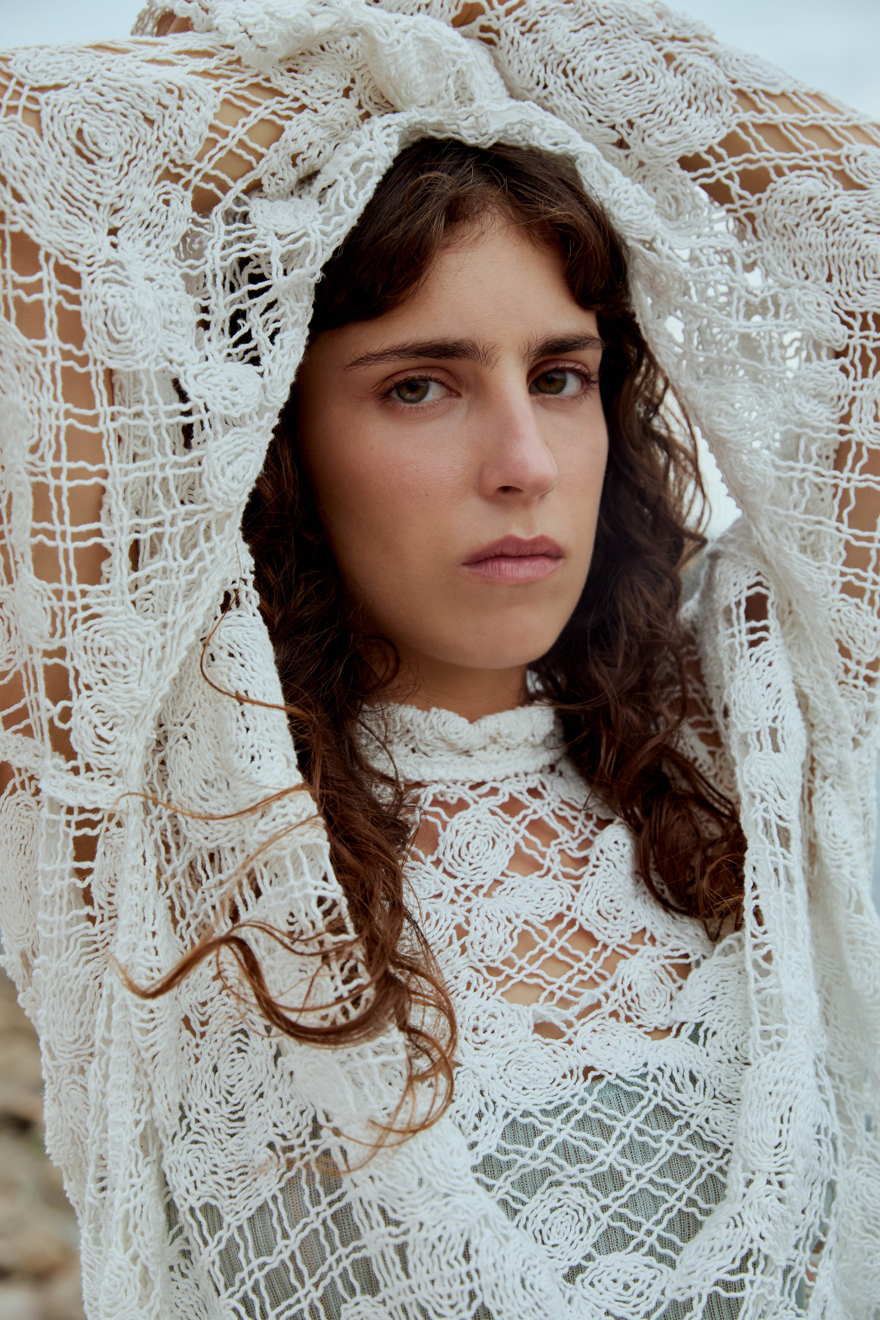 Woman with wavy hair wearing a lace garment, arms raised above her head, gazing seriously at the camera.
