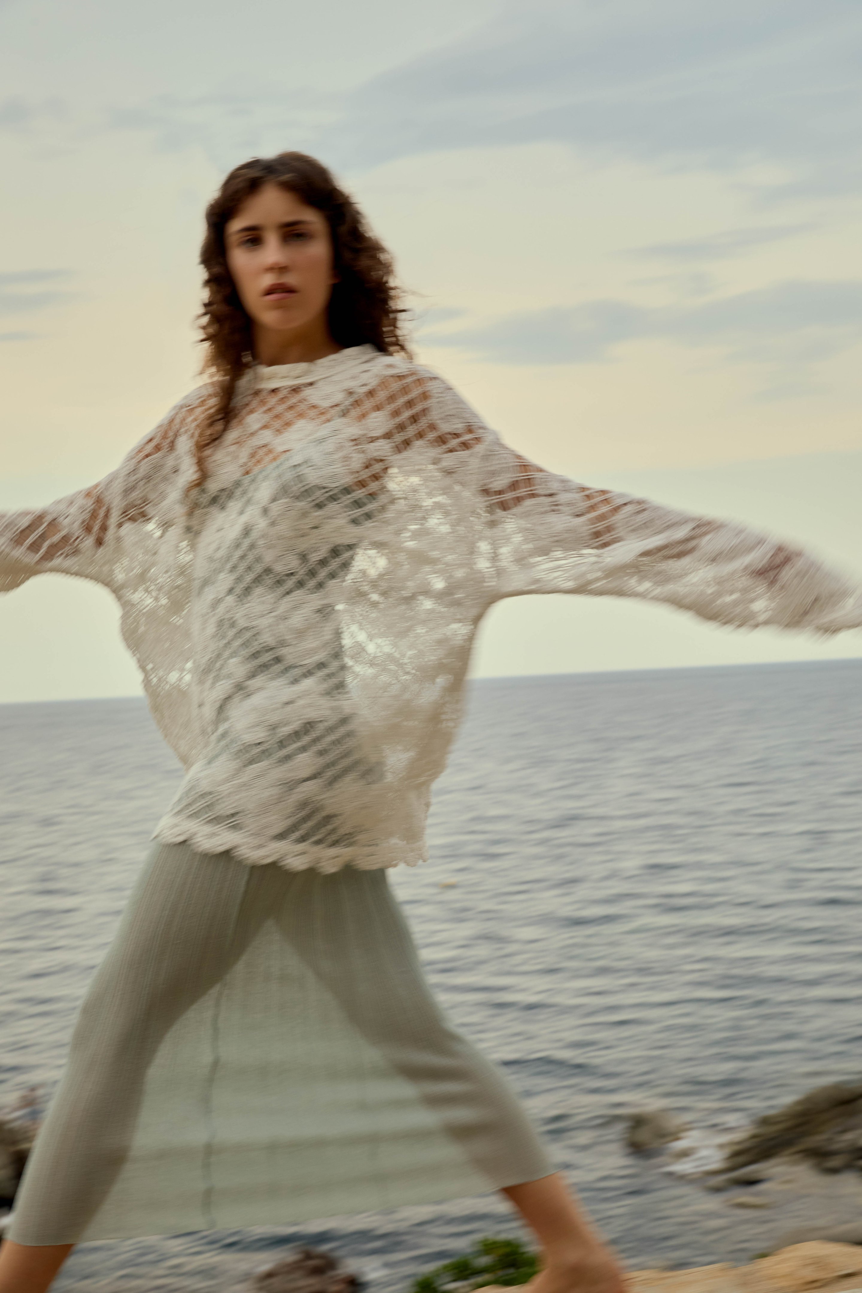 Frau trägt transparenten, weißen Spitzenponcho über einem mintfarbenen Outfit, geht am Meer entlang bei Sonnenuntergang