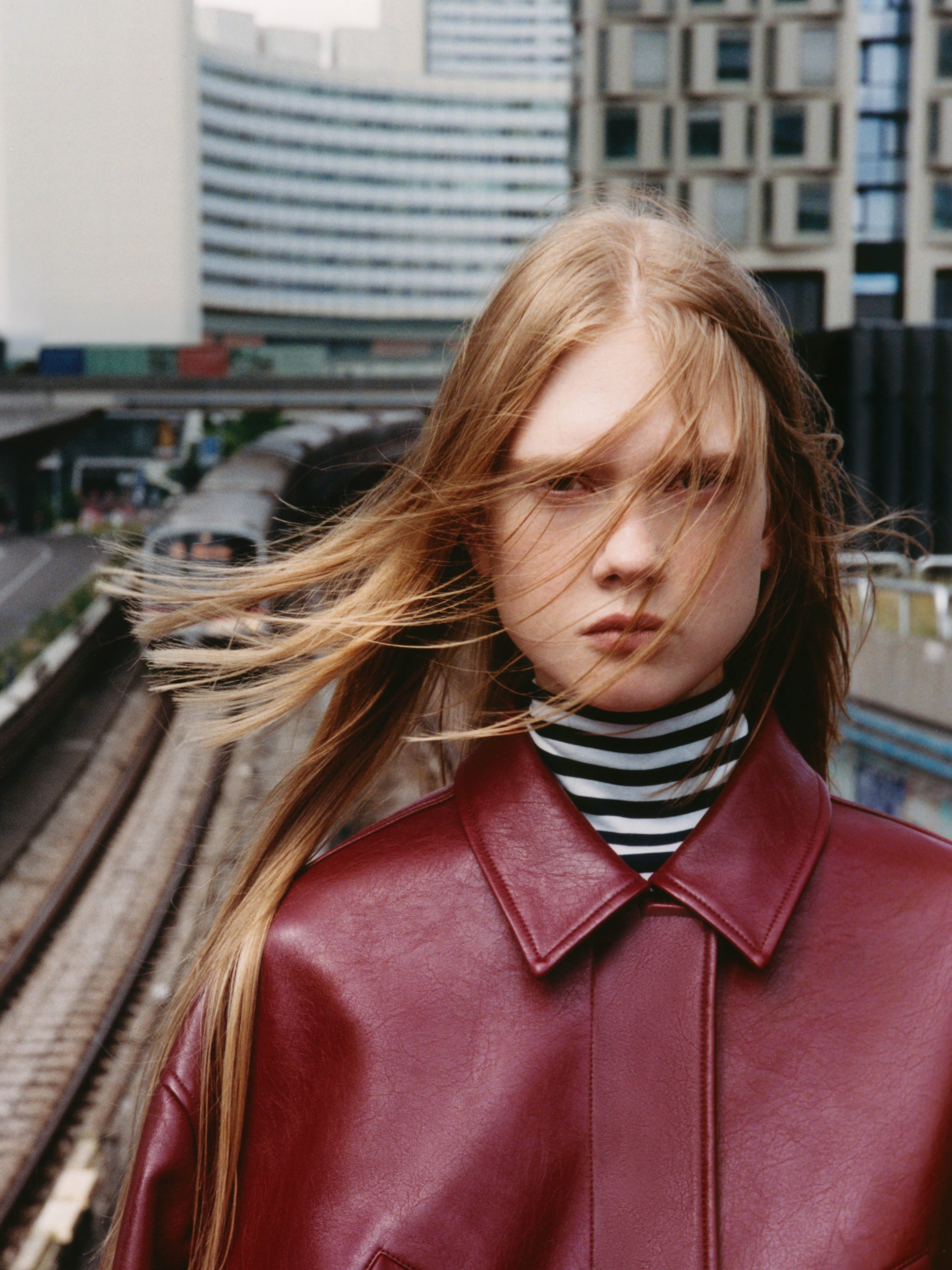 A person with long hair stands on a railway platform, wearing a red leather jacket and striped turtleneck, with buildings in the background.