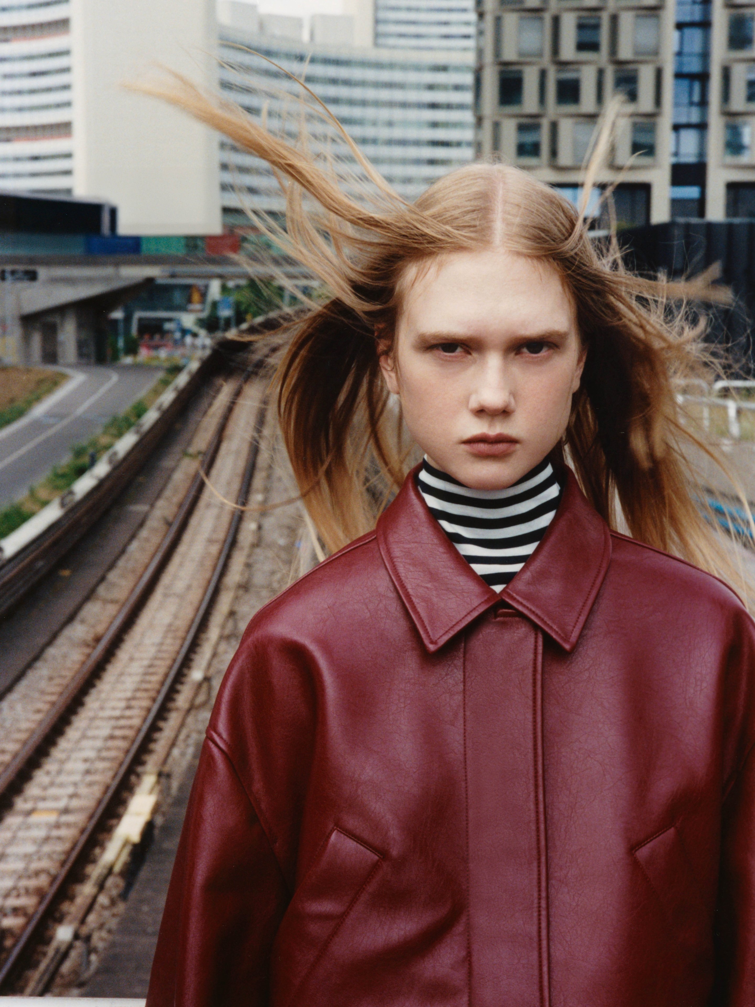 Person with long hair blowing in the wind, wearing a red leather jacket and striped turtleneck, standing near railway tracks in an urban setting.