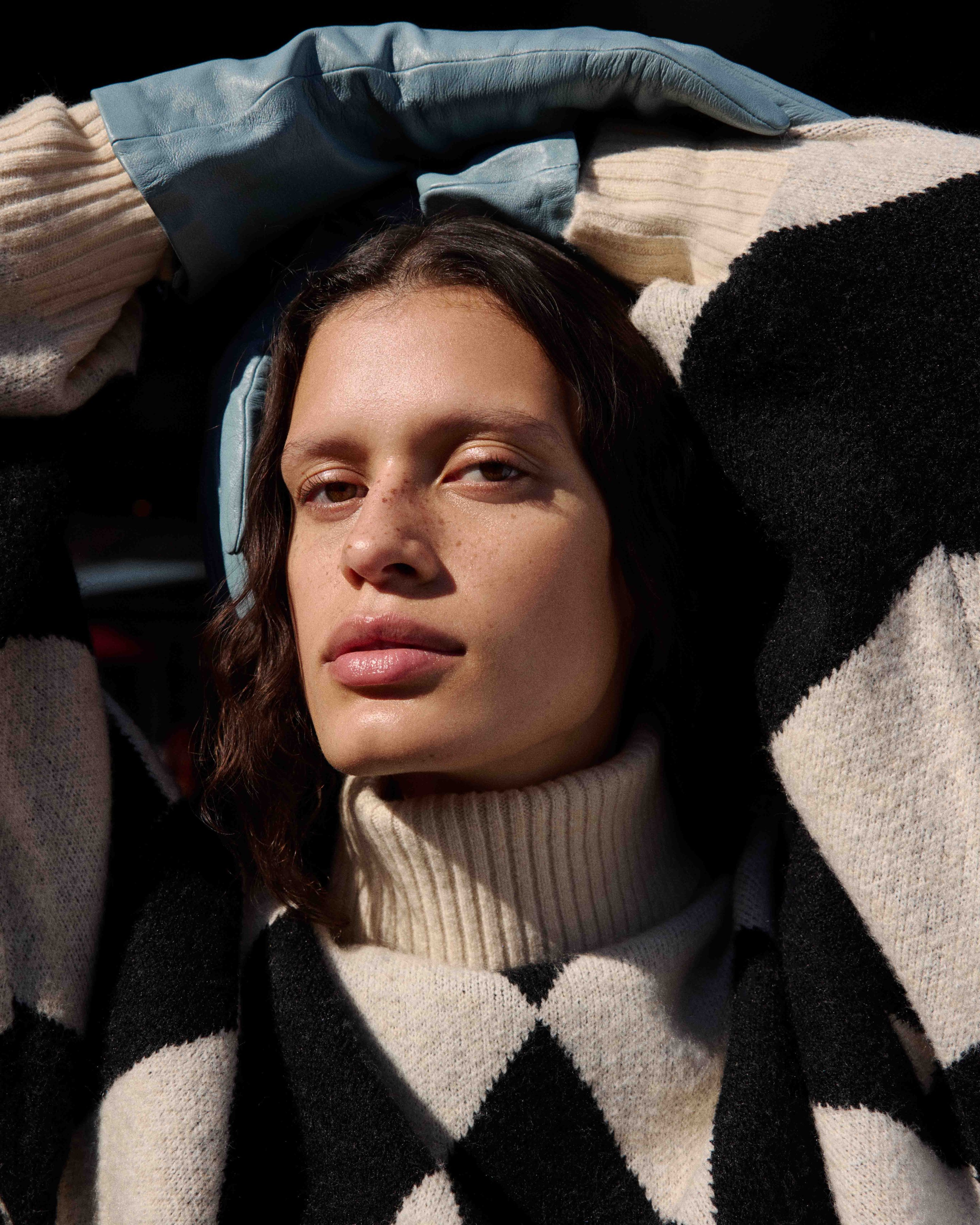 Person wearing a checkered sweater and blue jacket, with arms raised and hands resting on head, gazing into the distance.