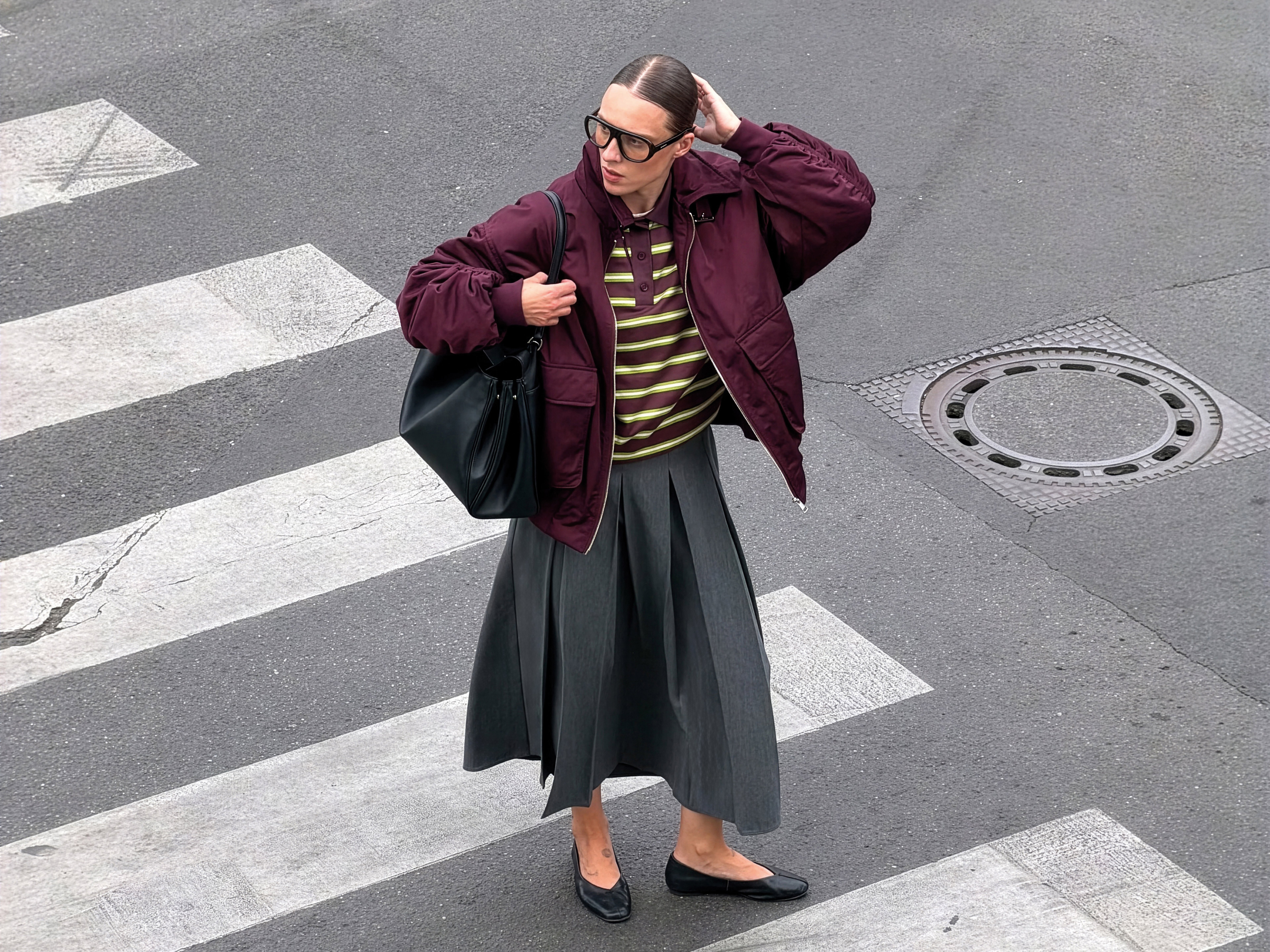 A person with sunglasses walks across a street in a burgundy jacket, striped shirt, gray skirt, and black flats, holding a large black bag.