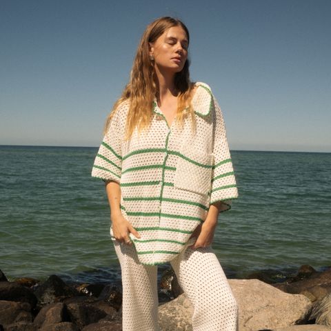 Woman in a patterned outfit standing by the sea on a sunny day, with rocks and the ocean in the background.