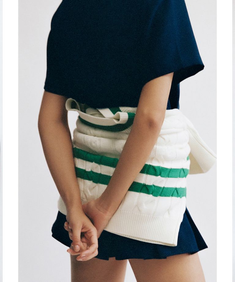 A person in a navy outfit with hands behind their back, wearing a white sweater around their waist with green stripes.