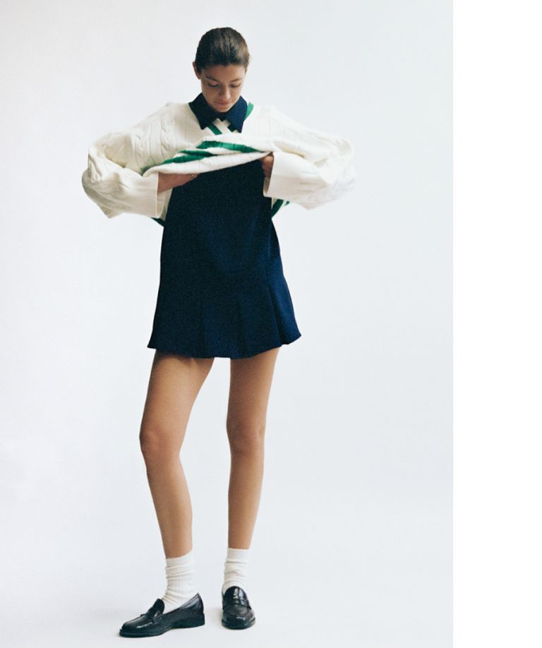 Person wearing a navy dress, white oversized sweater, white socks, and black loafers, standing against a plain white background.