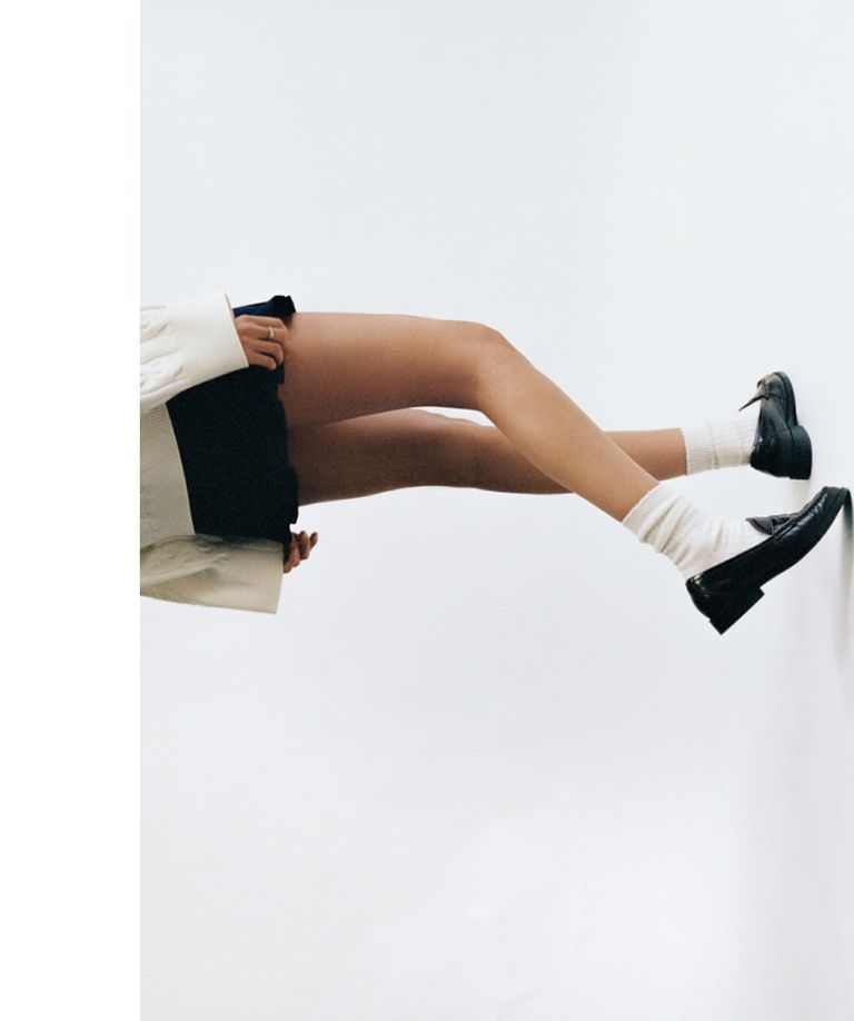 Person leaning sideways with legs extended, wearing a beige coat, black shorts, white socks, and black shoes against a white background.